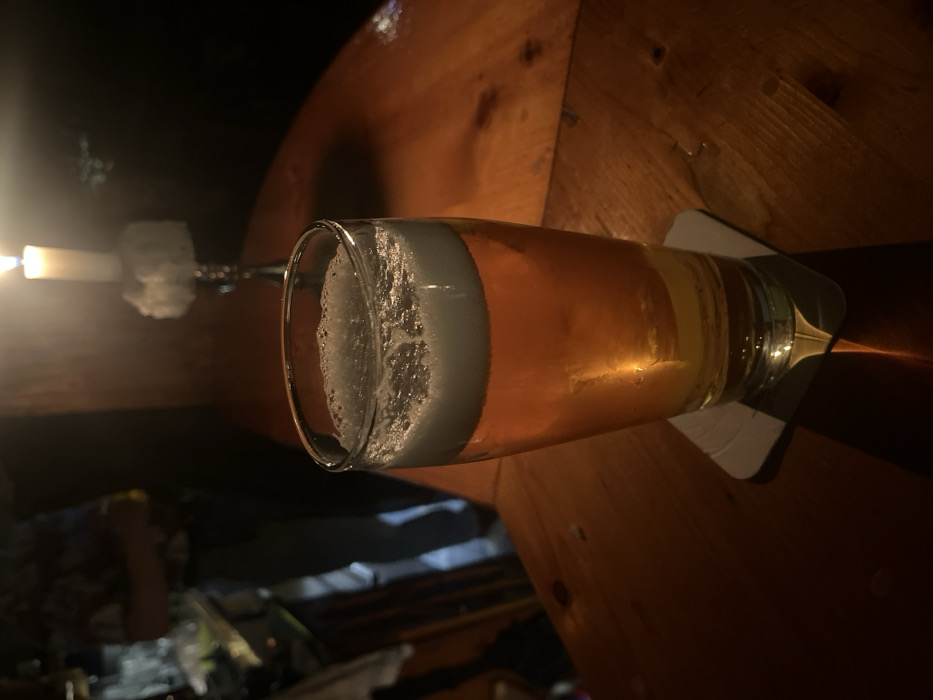 This media file presents a close-up, horizontally oriented view of a glass of beer resting on a wooden table, likely in a pub or bar setting in Bamberg, Germany. The beer, appearing light golden or amber with a substantial head of white foam, sits on a light-colored coaster. The glass reflects ambient light, highlighting the liquid's clarity.
To the left, a single lit candle in an ornate, possibly metal, candle holder casts a warm, soft glow across the scene, creating prominent shadows and illuminating the beer and table surface. The wooden table exhibits a rich, warm brown tone with visible grain, suggesting a rustic or traditional ambiance. The background beyond the table is dark and out of focus, implying a dimly lit environment, characteristic of an evening or nighttime setting.
The overall atmosphere conveyed is one of quiet relaxation and intimacy. No people are visible in the frame, and there is no discernible text. The focus remains on the drink and the subtle interplay of light and shadow provided by the candle.