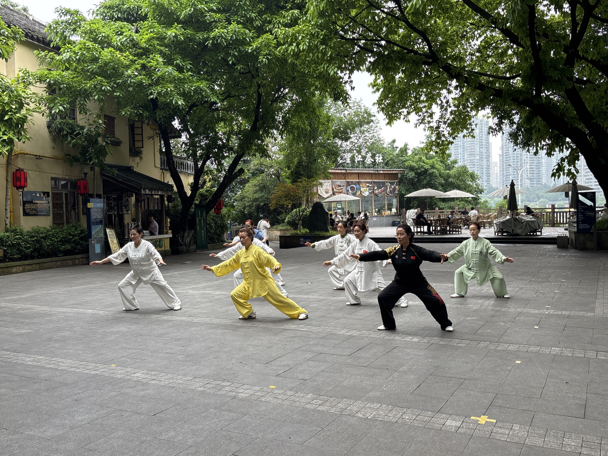 A group of seven women is practicing Tai Chi in Yuzhong District, Chongqing, China. They are dressed in traditional Tai Chi uniforms in a variety of colors including white, yellow, green, and black. The woman in the center is wearing a black uniform with a red Chinese flag patch on the shoulder. They are standing on a paved plaza made of gray rectangular pavers with a border of smaller, square pavers.
Behind the women, there are several buildings and trees. To the left, there is a two-story beige building with red lanterns hanging outside and to the right, there is an outdoor restaurant area with tables and umbrellas. In the background, there are modern high-rise buildings, suggesting an urban environment. The scene is set during the daytime under an overcast sky.