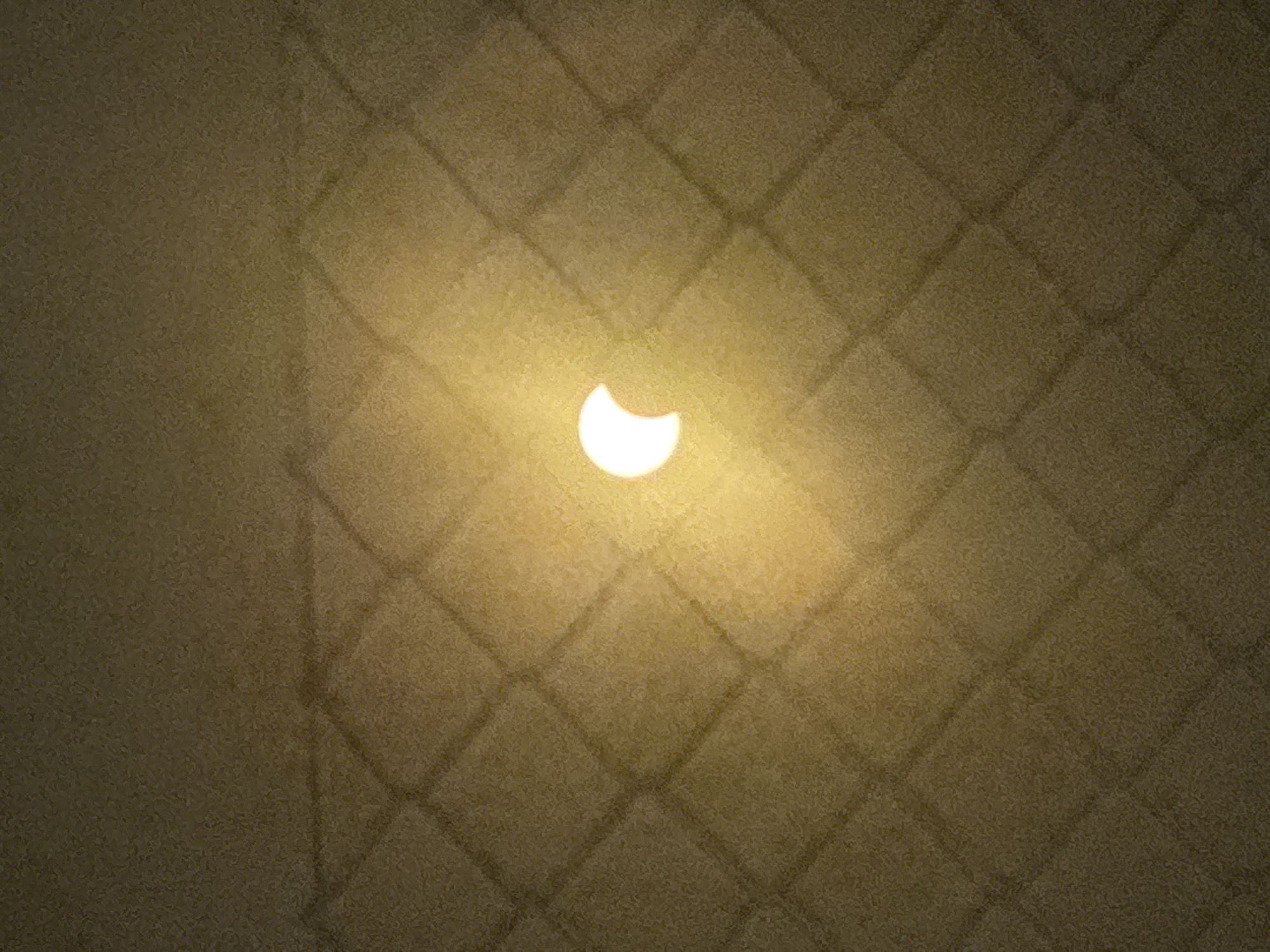 The image is a close-up, angled shot of a partially eclipsed sun projected onto a textured, tiled surface, likely a wall or pavement. The projection creates a diamond-shaped pattern of light and shadow, with the crescent of the eclipsed sun illuminated at the center of a diamond. The surface appears to be made of light brown or beige tiles, possibly stone or ceramic, with a rough texture. The lighting is warm and casts soft shadows. The image does not contain any people, specific landmarks, or identifiable elements that would place it in Calp, Spain. The scene depicts a natural phenomenon (solar eclipse) as observed through a projection method.