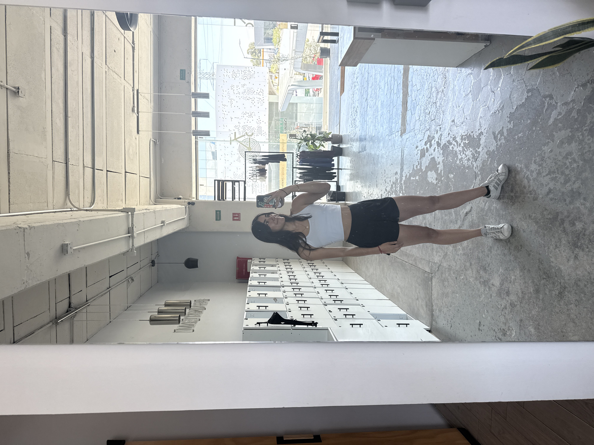 The image captures a young woman taking a mirror selfie in a modern, industrial-style interior space. The woman, with long dark hair, is dressed in a white crop top, black shorts, and white sneakers. She holds a smartphone to photograph her reflection, displaying a slight smile.
The setting features light gray concrete floors, partly adorned with a textured hexagonal tile pattern. The walls are constructed from light-colored concrete blocks, with exposed electrical conduits running along them and the ceiling. In the reflection, a long row of white lockers with black handles is visible, alongside a red box mounted on the wall above them. Behind the woman, a black clothing rack displays various garments, and a potted plant is also visible. Modern light fixtures, including cylindrical and bell-shaped designs, hang from the ceiling.
Large windows provide a bright view of an urban outdoor scene. Through the glass, other buildings, trees, and a street are discernible. A distinctive white architectural or decorative structure, perforated with numerous small holes, is prominent in the outdoor view.
The bright ambient light indicates it is a sunny daytime. Visible text includes a green "SALIDA DE EMERGENCIA" (Emergency Exit) sign, appearing twice due to the mirror's reflection. The words "STUDIO" and "ROWBO" are also partially visible as reflections on the clothing rack. The overall atmosphere of the space is clean and contemporary. While the location context is Puebla, Mexico, this is not visually verifiable from the image.