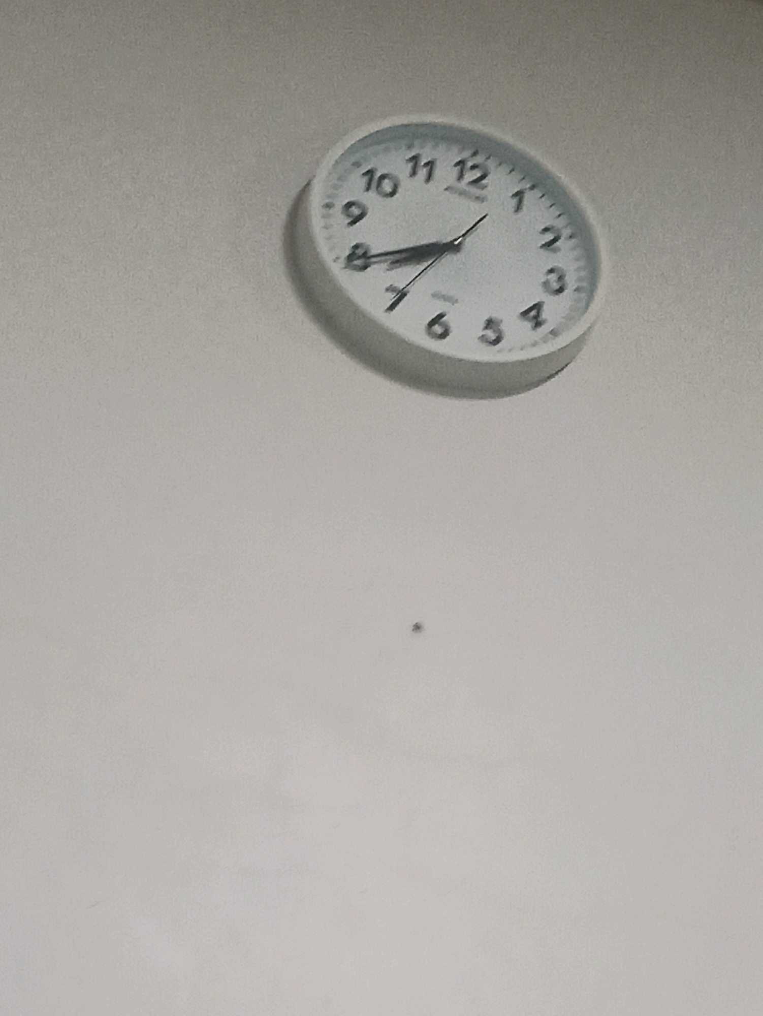 A close-up, slightly blurry, overhead shot shows a white analog clock on a light gray wall. The clock has black Arabic numerals and black hands. The hour hand points between 9 and 10, and the minute hand points between 1 and 2. A small, dark speck is visible on the wall below and to the left of the clock. The lighting suggests an indoor environment.