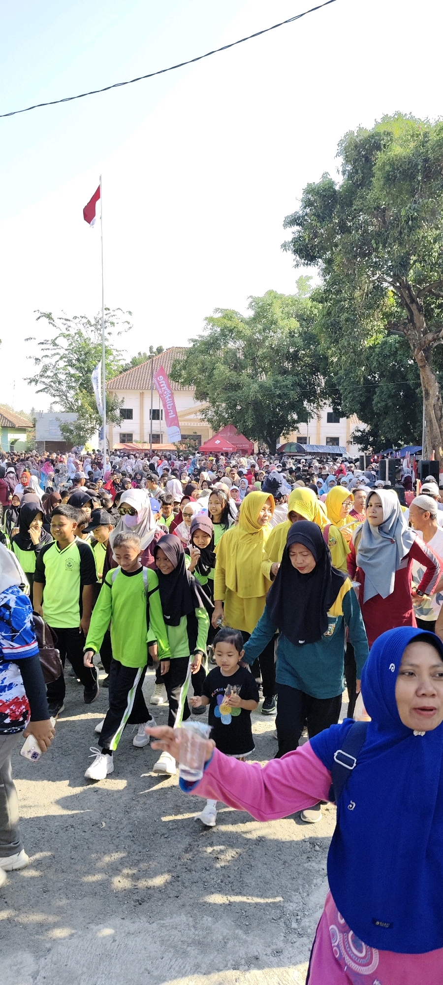 The image depicts a crowded outdoor scene in Sulang, Indonesia, likely during a community event or procession. A large group of people, including men, women, and children, are walking together. Many individuals are wearing headscarves and traditional Islamic clothing. Some people are wearing bright colored athletic wear, suggesting a possible fun run or walk. In the background, there are buildings and trees. A red and white flag is visible on a flagpole, which could be the Indonesian flag, indicating a patriotic or nationalistic event. The ground is paved with what appears to be asphalt or concrete. The overall atmosphere seems lively and communal.