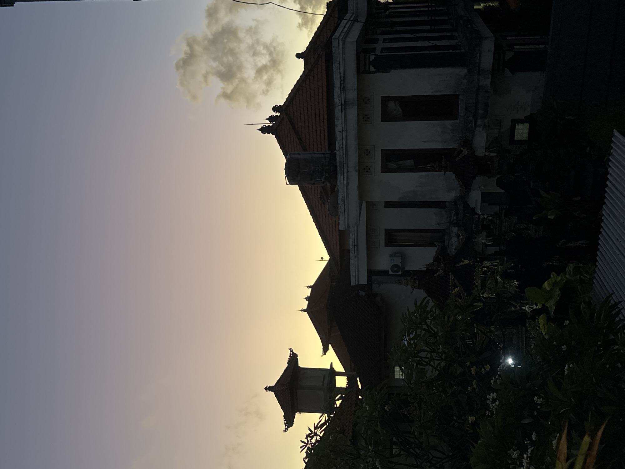 The image displays an outdoor scene at either sunrise or sunset, characterized by a soft, warm gradient in the sky transitioning from golden-yellow near the horizon to a muted purple-blue overhead. Silhouetted, dark clouds are visible in the upper portion of the sky.
The main subjects are traditional Balinese-style buildings, consistent with the Kuta, Indonesia location. The prominent building in the center features a multi-tiered, reddish-brown tiled roof with decorative finials. Its light-colored walls appear weathered, and dark, empty window openings are visible. A black water tank rests on this building's roof, alongside what appears to be an air conditioning unit and a partially visible satellite dish. To the left, a smaller, ornate temple-like structure with a similar tiered roof is silhouetted against the bright sky.
Lush, dark green tropical foliage, including bushes and trees with some white flowers, fills the foreground and right side of the image, appearing almost black due to the low light. A bright, circular light source, possibly a lamp, glows faintly through the dense greenery on the lower right. A corrugated metal fence is partially visible on the far right.
The scene is tranquil and devoid of people or active events. The low sun angle creates deep shadows, accentuating the architectural details and vegetation. No visible text is present in the image.