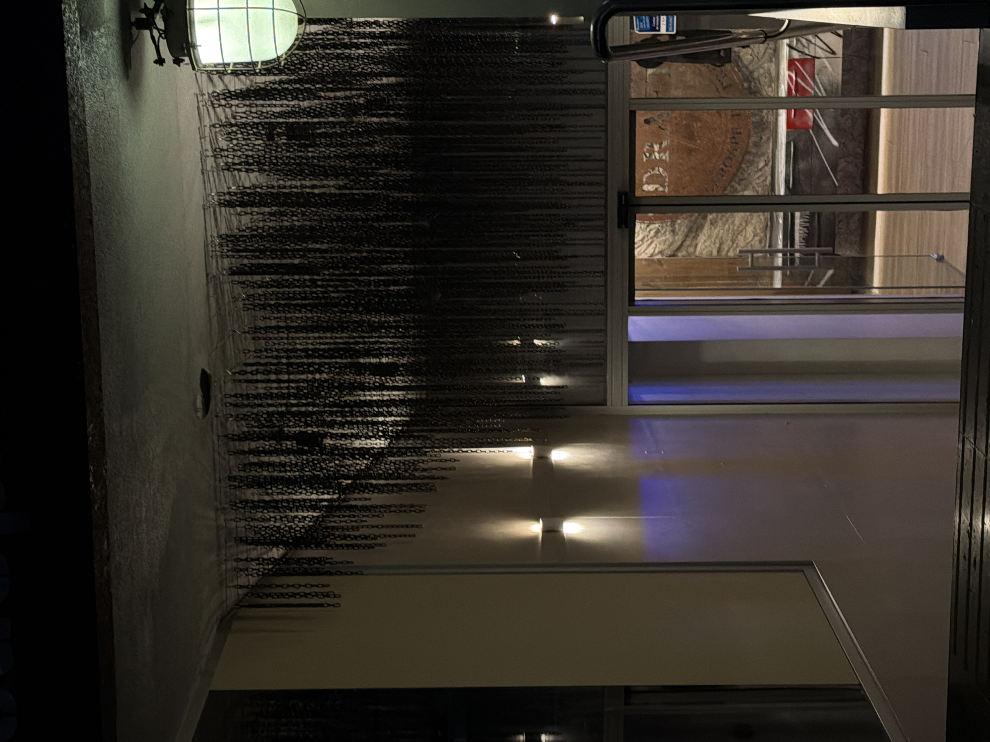 The image shows an exterior shot of a building at night. A green-tinted light fixture is attached to the wall. Adjacent to the light fixture, a curtain of chains hangs down. Further along the wall, there are some outdoor lights casting light onto the wall. To the right, there is a window, which reveals an interior space that is out of focus, but includes lettering on a round object and a red object.