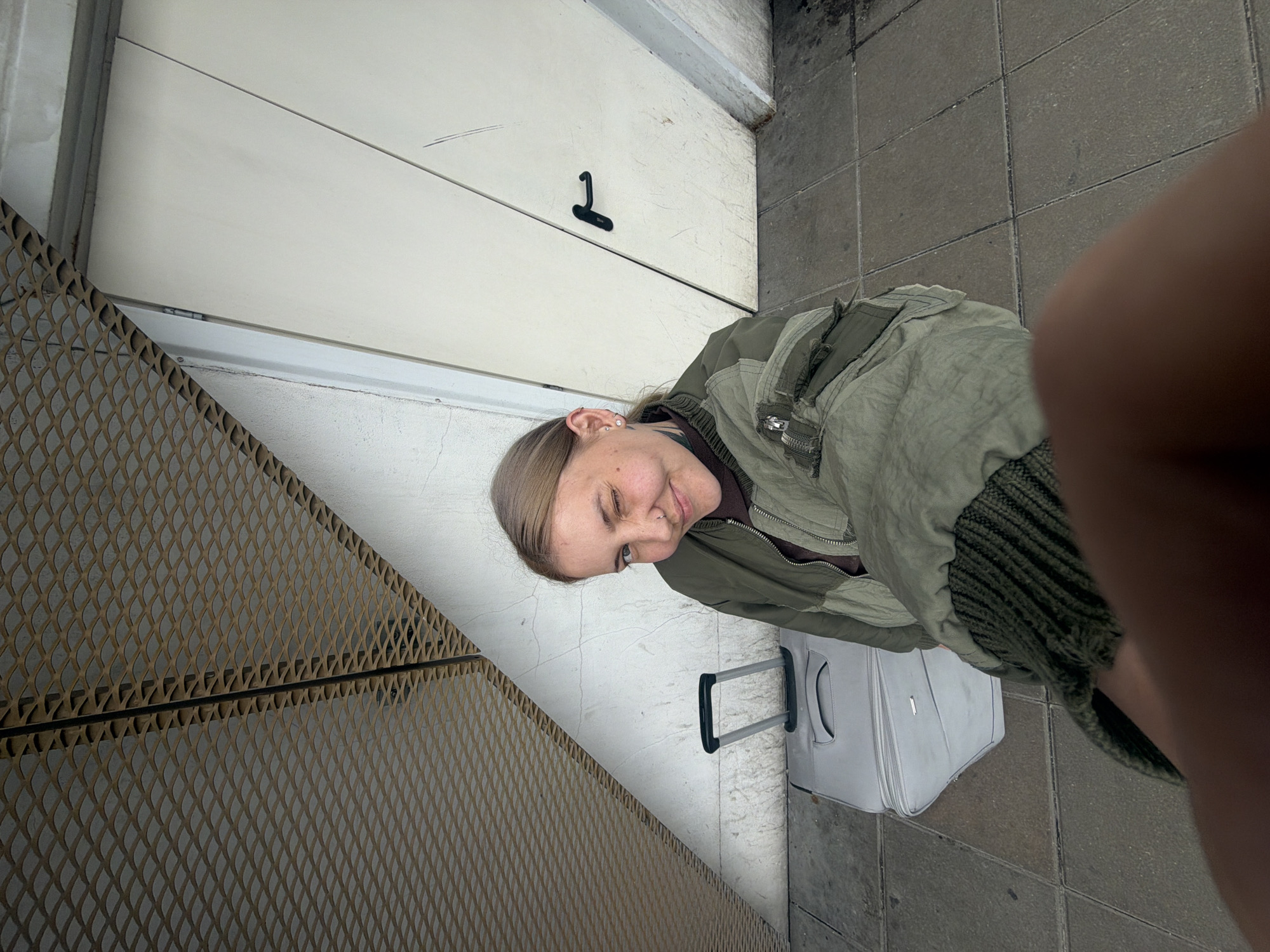 A young, light-skinned woman with blonde hair is standing in an urban environment, partially obscuring the camera's view. She is wearing an olive-green jacket with a green, knitted collar. She has a small carry-on suitcase with a silver handle beside her. The ground consists of gray tiled pavement. To the left is a textured, metal mesh screen. Behind her is a wall with a white door, marked with a black "1". The location is Khimki, Russia.