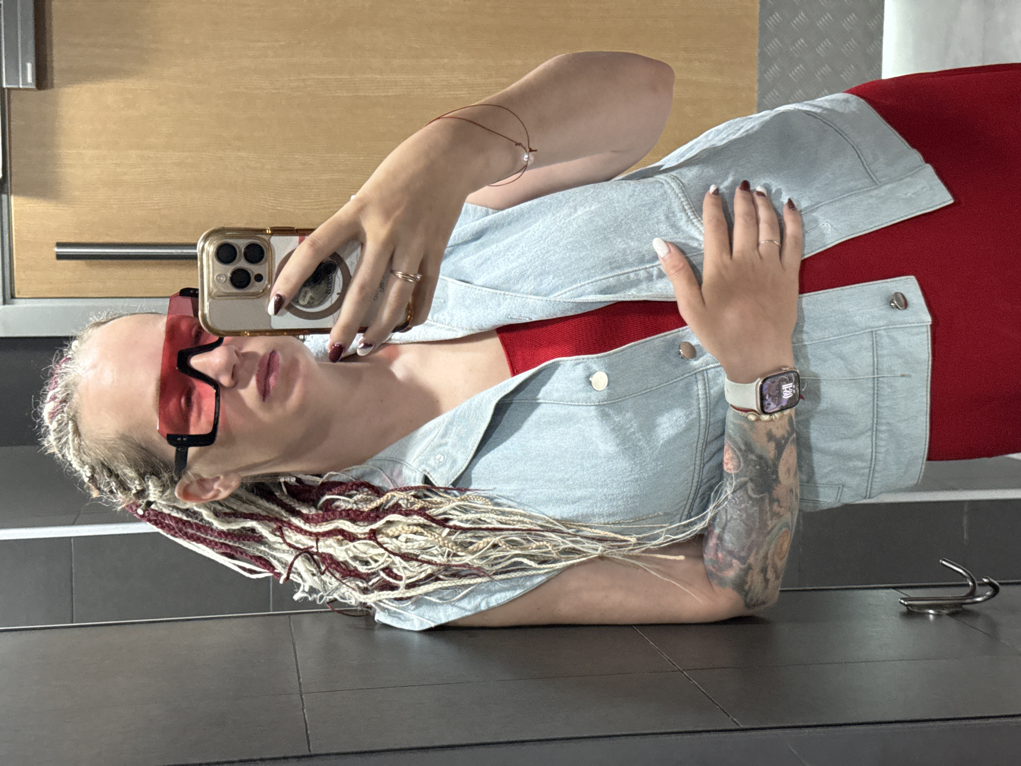 A woman with blonde and burgundy braids is taking a mirror selfie in a locker room. She is wearing a red dress, a light blue denim vest, and large red-tinted sunglasses. She has tattoos on her left arm and is wearing a smartwatch and rings. The background shows a grey tiled wall and a wooden door with a metal handle. The image is taken in Sochi, Russia.