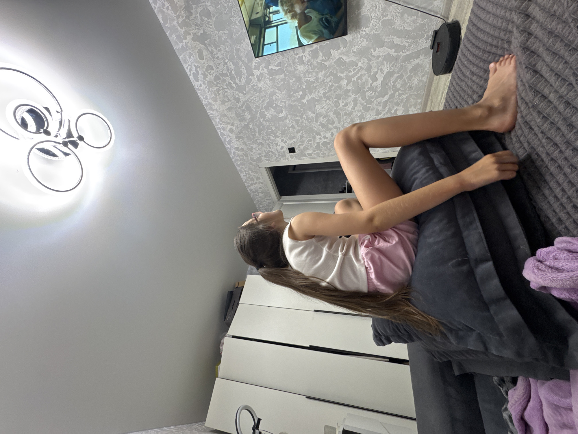 A young girl with long brown hair is lying on a grey upholstered surface, possibly a bed or sofa. She is wearing a white tank top and pink shorts. Her body is oriented vertically in the frame, with her head at the top and feet towards the bottom. Her right leg is bent at the knee, and her right arm is extended, resting on the surface. Her left arm is bent, and her hand is near her chest.
In the background, a modern ceiling light fixture with multiple circular elements is visible on the left. On the right, a television screen shows a scene with people, and above it, a wall with textured wallpaper. A small, round, black robot vacuum cleaner is on the floor near the edge of the upholstered surface. In the lower portion of the frame, a white dresser with dark handles is partially visible.
The scene appears to be indoors, in a bedroom or living room. The lighting suggests it could be daytime, as there is artificial light from the ceiling fixture and the television screen. There are no obvious cues about the weather. The overall mood appears relaxed, with the girl seemingly engaged with the television. No visible text is present in the image.