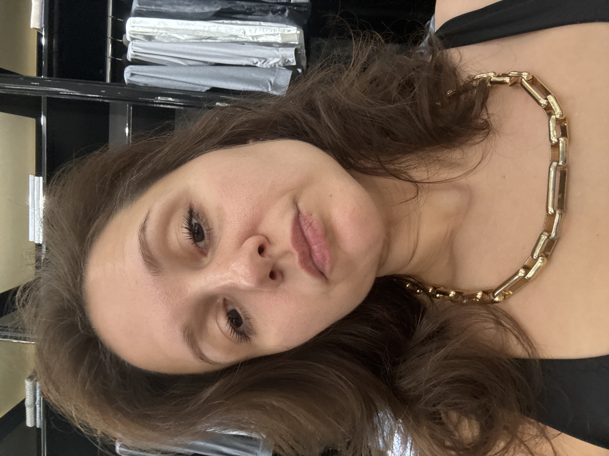 A woman with brown hair and a gold chain necklace is pictured indoors. Behind her is a black shelving unit with stacks of gray and white items. The location is not identifiable as Dubai, United Arab Emirates, from the visual information provided.
