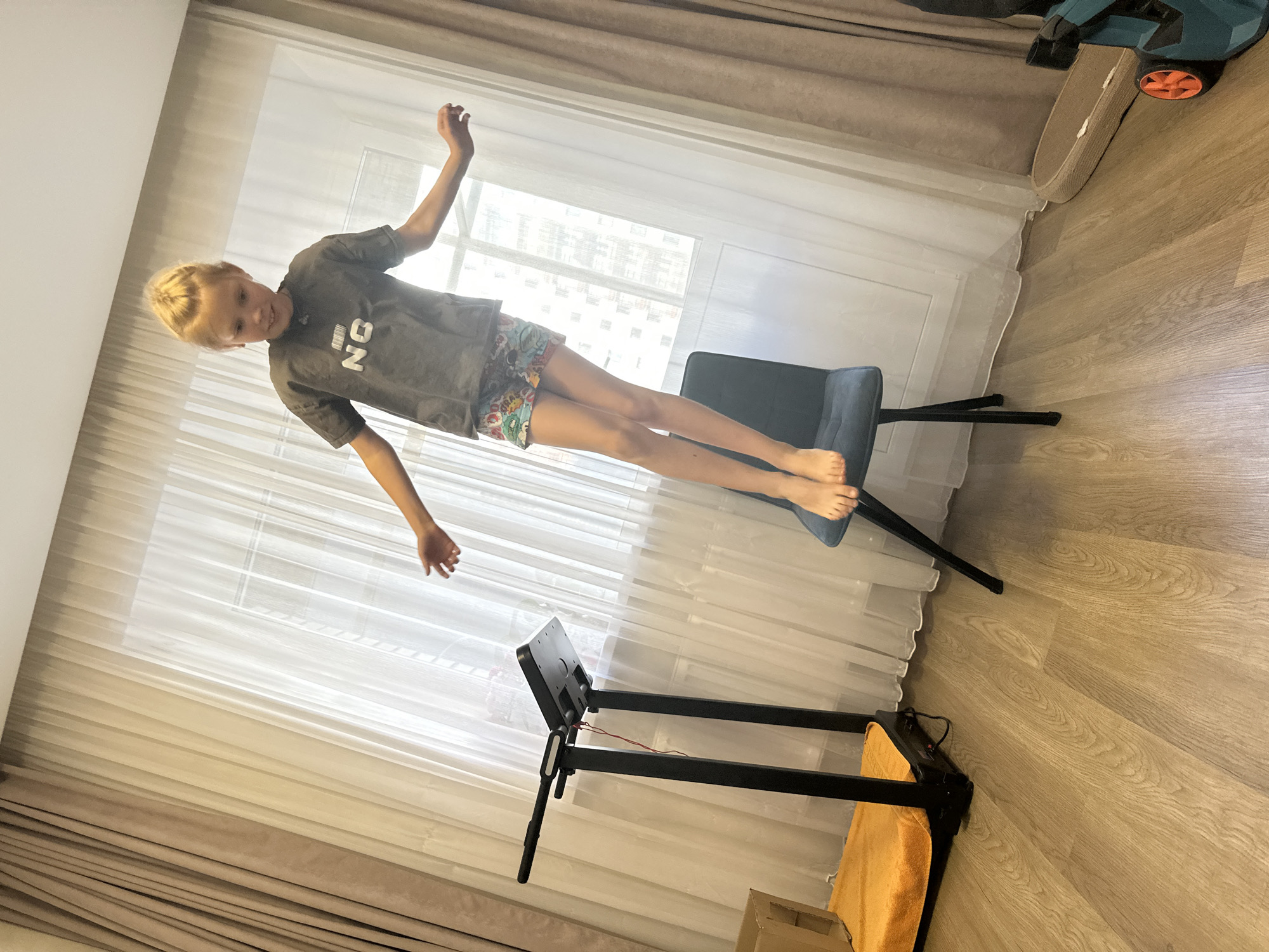 A young boy is playfully balancing on a dark blue chair, his body angled as if he's in mid-air. He's wearing a grey t-shirt with the letters "NO" printed in a blocky, barcode-like font and colorful patterned shorts. His blonde hair is short and styled upwards. His arms are outstretched, and one hand is reaching towards the ceiling. His legs are extended, and his feet are bare.
The boy is in an indoor setting with large windows covered by sheer white curtains and heavier beige curtains. The room has light wooden flooring. In the background, a treadmill with a black frame and an orange mat is visible. The lighting suggests it might be daytime, possibly late morning or early afternoon, with sunlight streaming through the window. The overall atmosphere appears playful and energetic, with the boy's pose suggesting a moment of fun or a staged activity. The location context is Abakan, Russia.