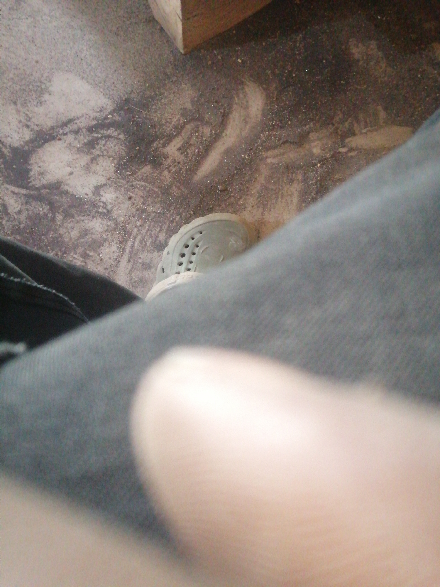 The image depicts a close-up, low-angle shot of a person's leg and foot wearing a white perforated shoe, possibly a clog or Croc-style sandal. The foot is resting on a textured, dusty surface that appears to be the ground, with some debris and what looks like dirt or dust patterns. A portion of dark-colored fabric, likely pants or jeans, is visible to the left of the foot, and a soft, out-of-focus area, potentially a finger or thumb, is in the foreground. In the upper right corner, a wooden object, possibly a piece of lumber or furniture, is partially visible. The overall scene suggests an indoor or outdoor area with a rough, unfinished floor.