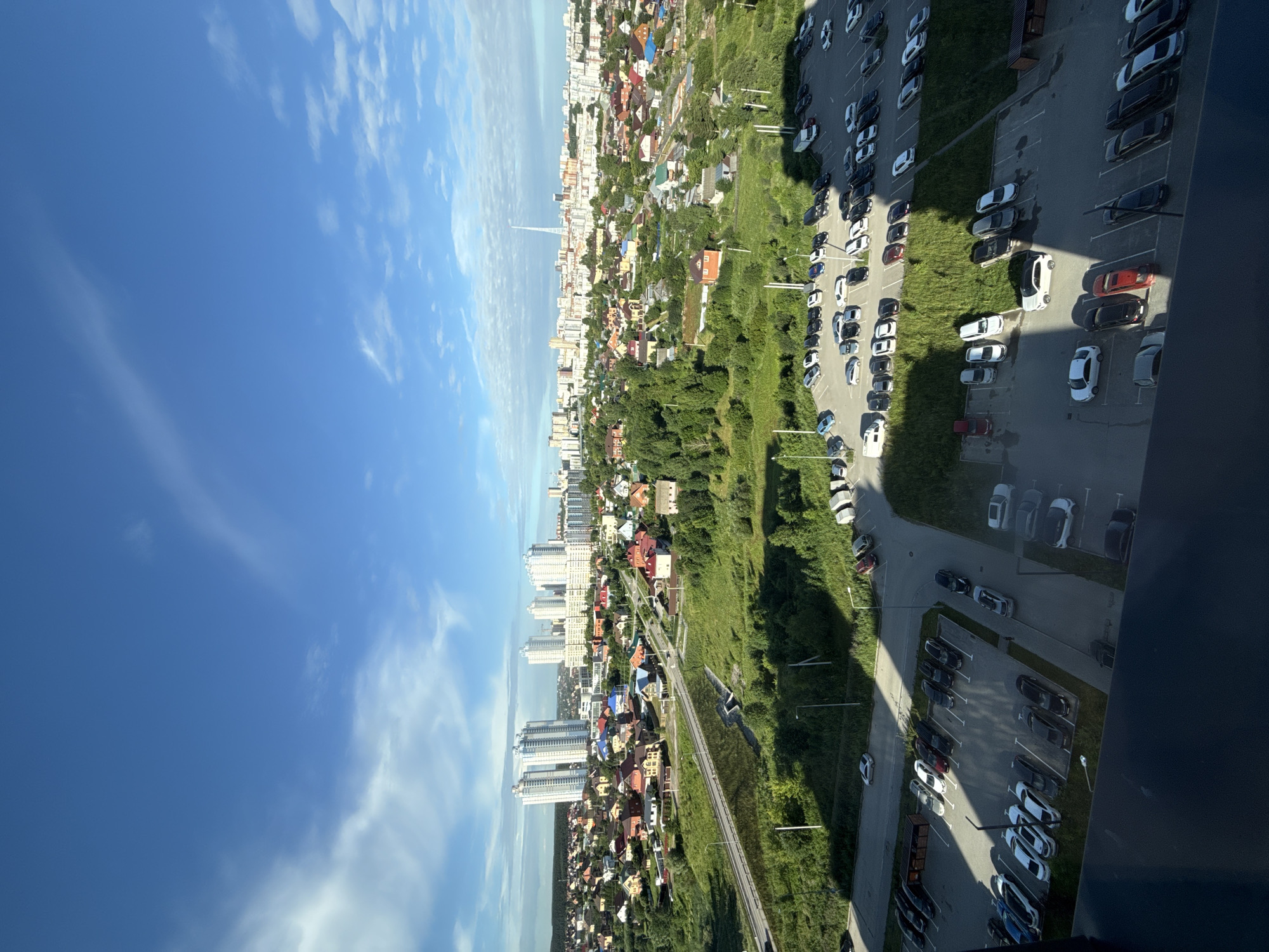 This is a photograph taken from a high vantage point, likely a building, looking out over a mixed-use urban and suburban landscape. The sky is a vibrant blue with scattered, wispy white clouds, suggesting a clear day.
In the foreground, a dark, reflective surface, possibly a window or balcony railing, partially obstructs the view. Below this, a large, paved parking lot is filled with numerous cars of various makes, models, and colors. The cars are parked in organized rows, with some spaces empty. A few small trash bins are also visible in the parking lot. The parking lot is bordered by green grass and trees.
Beyond the parking lot, a section of road is visible, with a few cars driving on it. This road leads into a densely populated residential area characterized by numerous houses with pitched roofs, painted in a variety of colors including red, yellow, and brown. Interspersed among the houses are trees and green spaces.
Further in the distance, a cluster of modern, multi-story apartment buildings stands out, appearing tall and white. Beyond these buildings, the cityscape continues with more structures, suggesting a larger urban center. A tall, slender tower, possibly a communication or observation tower, is visible on the horizon. The overall impression is of a sunlit day in a developing area, where suburban housing transitions into a more urban environment.
The location is identified as the city of Perm, Russia. Specifically, the scene appears to depict the microdistricts of Novye Lyady or adjacent areas, given the characteristic mix of single-family homes, newer apartment complexes, and proximity to green spaces and infrastructure seen in the image. The presence of high-rise buildings and a mix of housing types is consistent with areas undergoing development and expansion in Perm.