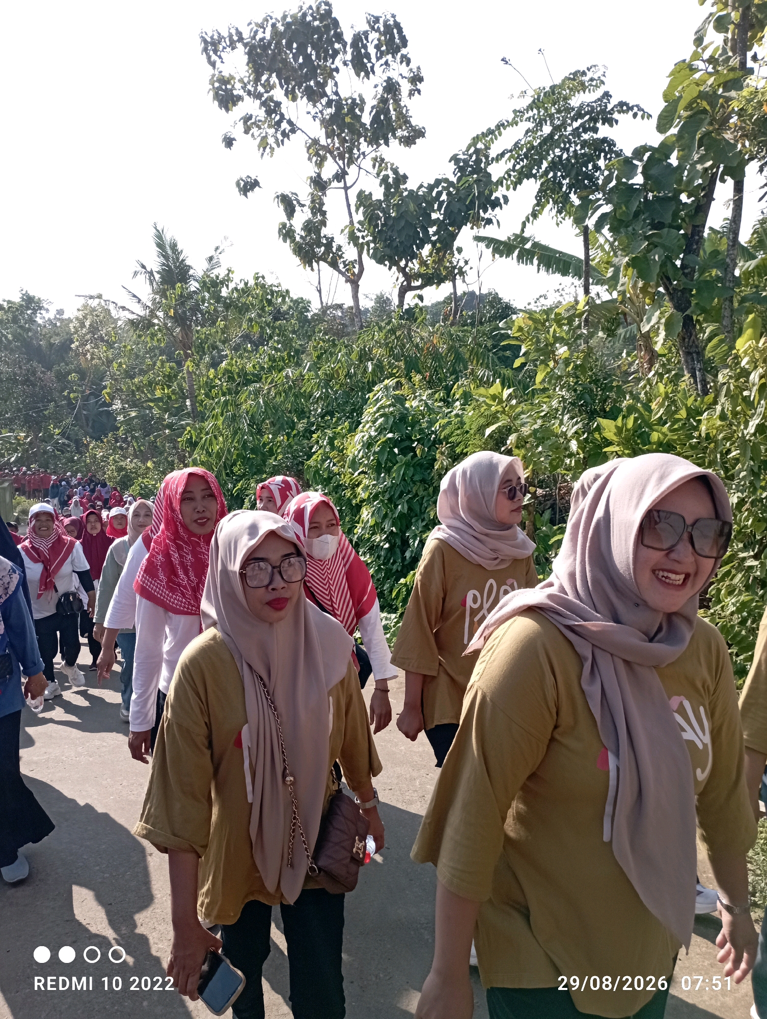 A group of women wearing headscarves are walking along a paved road in a rural, outdoor setting. They are mostly wearing brown or gold shirts. The woman in the foreground on the right is smiling widely and wearing large sunglasses and a light brown headscarf. The woman in the foreground on the left is wearing glasses and a light brown headscarf and looking forward. Behind them, many other women are walking in a line. The road is lined with lush green trees and vegetation. The sky is bright, indicating it is daytime. A date stamp in the bottom right corner reads "29/08/2026 07:51". Another stamp in the bottom left corner reads "REDMI 10 2022". The image appears to have been taken in Dukuhseti, Indonesia, as indicated by the text in the prompt.