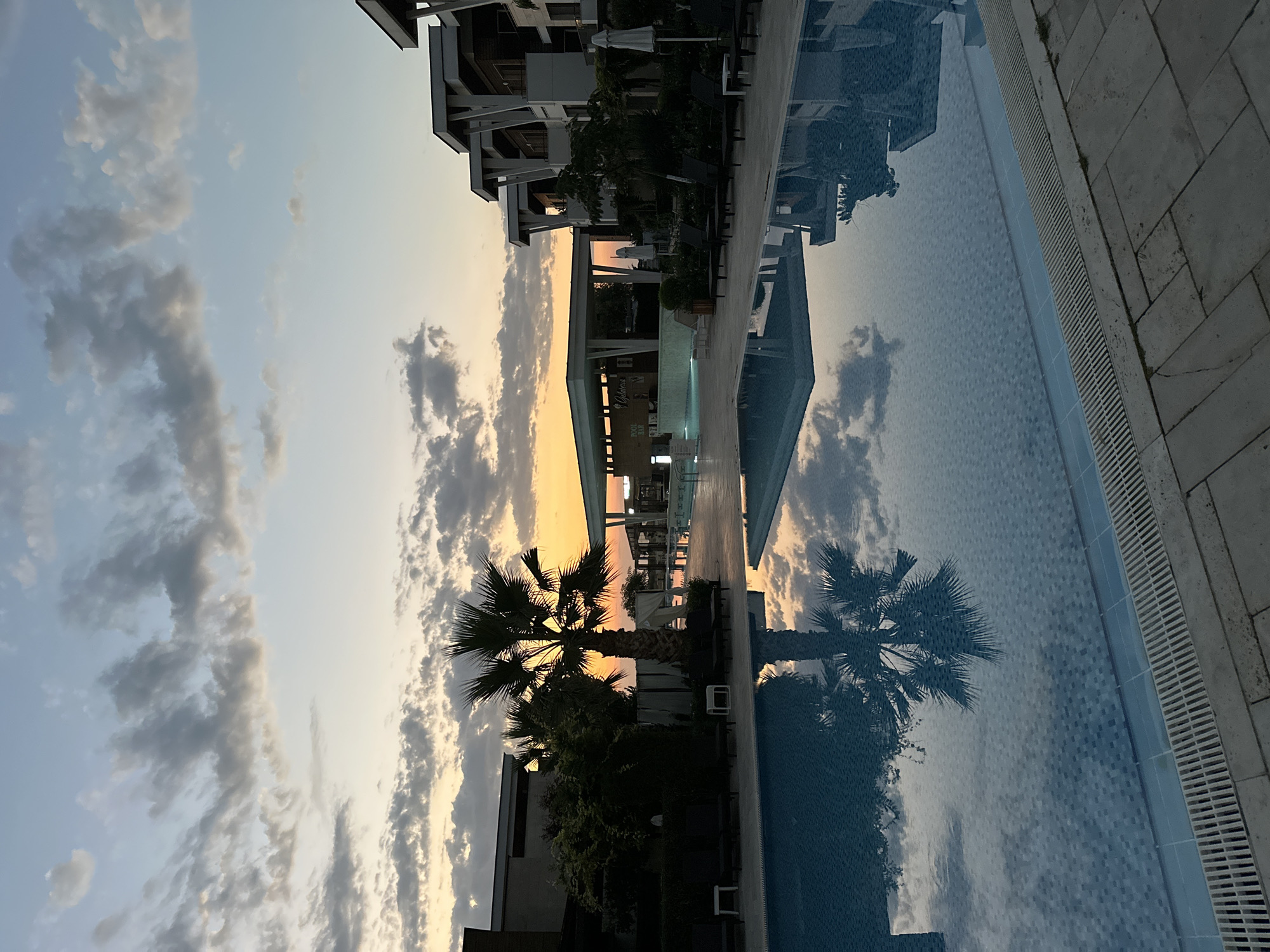 The image depicts a tranquil resort scene at sunset in Sozopol, Bulgaria. A large swimming pool dominates the foreground, its surface reflecting the pastel hues of the sky and the silhouette of a palm tree. In the background, modern resort buildings with clean lines and white architectural elements are visible. A bar area with seating and umbrellas suggests a place for relaxation and entertainment. The sky is adorned with scattered clouds, illuminated by the warm glow of the setting sun, creating a serene and picturesque atmosphere. The overall scene conveys a sense of peace and luxury associated with a holiday destination.