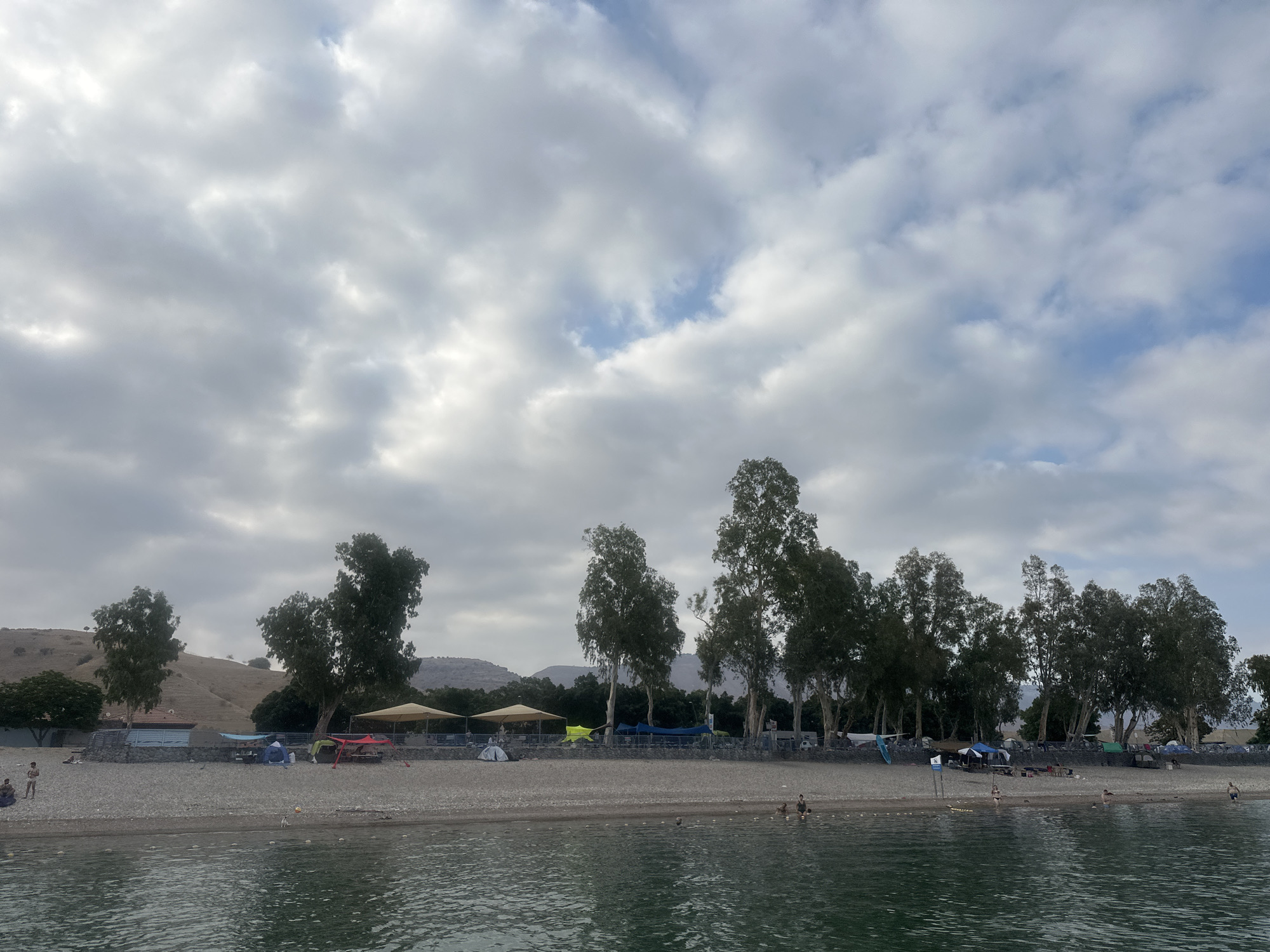 This image depicts a lakeside scene in Emek HaYarden Regional Council, Israel, likely on the Sea of Galilee. The foreground shows calm water with ripples, reflecting the sky. The midground features a pebbly beach with scattered people engaged in various activities. Several tents and canopies are set up, suggesting a camping or recreational area. Trees line the edge of the beach, providing shade. In the background, rolling hills are visible under a cloudy sky.