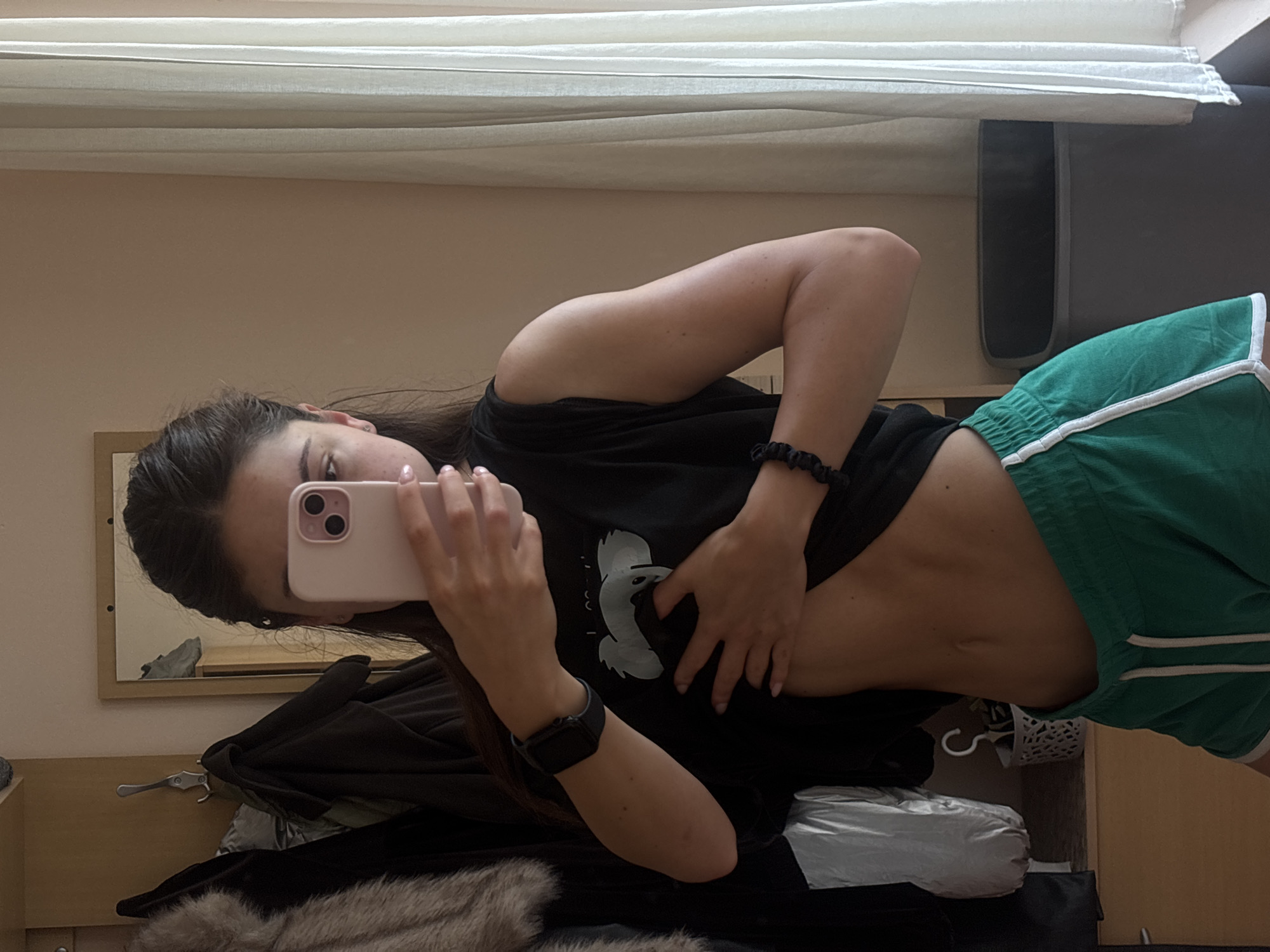 A young woman with dark hair pulled back is taking a mirror selfie in a room. She is wearing a black sleeveless shirt, which she has lifted to reveal her toned abdomen, and green shorts with white trim. She is holding a pink smartphone in her right hand, obscuring her face, and her left hand is on her stomach. A black smartwatch is visible on her left wrist, and a black scrunchie is on her right wrist. In the background, there is a closet with clothes hanging, a mirror reflecting a pair of shoes, a white basket with a hook, a trash can, and a window with white curtains. The overall impression is of a domestic setting. The image does not provide any explicit clues about the specific location within the city of Varna, Bulgaria.
