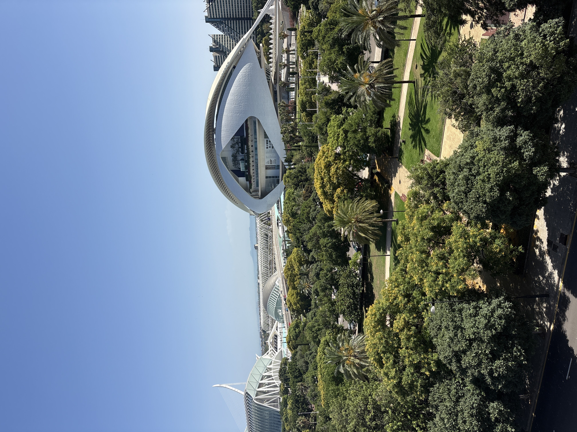 The image depicts a daytime, sunny scene in València, Spain, featuring a modern architectural complex surrounded by lush greenery. The primary subject is the striking architecture, characterized by a large, futuristic building with a distinctive white, curved facade, resembling an eye or a shell. This building is part of a larger complex that includes other contemporary structures with angular designs and glass elements, as well as bridges and elevated walkways.
The setting is a vibrant park or garden area, filled with mature trees, including palm trees, and well-maintained lawns. Pathways wind through the park, some paved with stone and others with lighter colored materials. Sunlight casts distinct shadows from the trees and palm fronds onto the grass and pathways, indicating a bright, sunny day. There are no people clearly visible in the image, suggesting a moment of quiet in the public space.
In the background, the urban environment of València is visible, with tall modern buildings and a glimpse of what appears to be the sea or a large body of water in the distance. The sky is a clear, bright blue. The overall impression is one of a contemporary urban oasis, blending modern architecture with nature. The image is oriented vertically, suggesting it was taken from an elevated viewpoint. No discernible text is visible in the image.