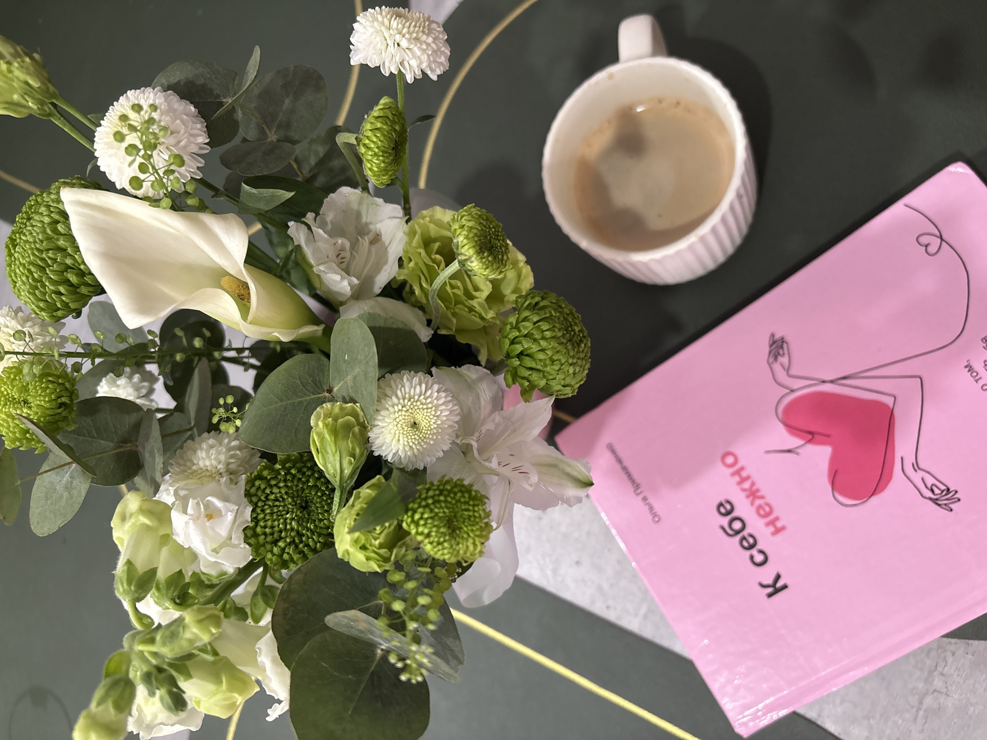 A top-down shot shows a bouquet of flowers, a white mug with coffee, and a pink book on a dark green surface with a gold line. The bouquet consists of white calla lilies, white button flowers, green pompom flowers, and eucalyptus leaves. The coffee mug is white with vertical ridges and has a handle. The pink book has text in Russian and an illustration of a body with a heart. The image was taken in Chaikovsky, Russia.
