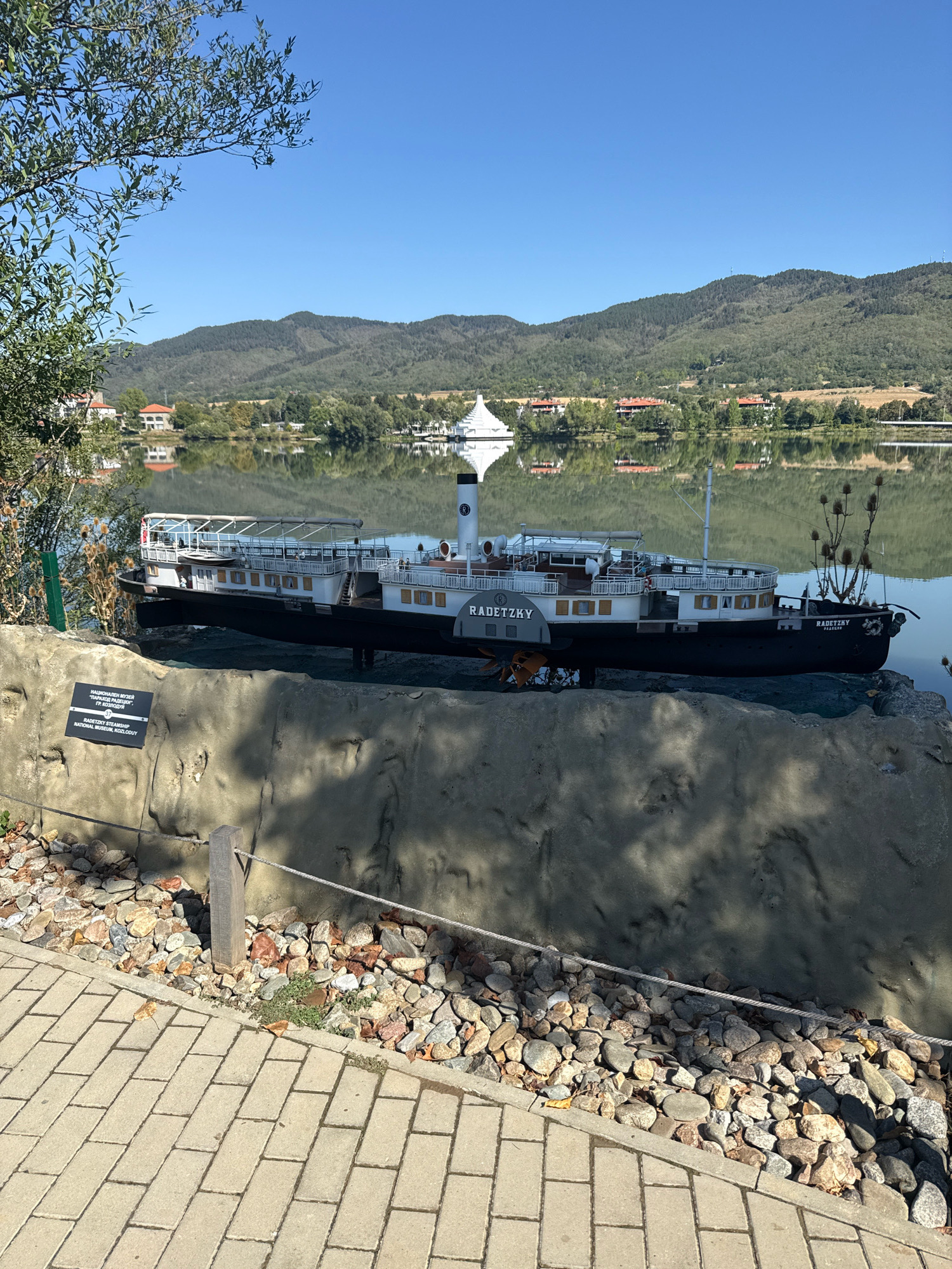 A model of the steamboat "Radetzky" is displayed on a rocky embankment overlooking a tranquil lake. The steamboat is black and white with multiple decks and a prominent smokestack. The name "RADETZKY" is visible on the side of the vessel. A sign in Bulgarian and English identifies it as the "RADETZKY STEAMSHIP NATIONAL MUSEUM, KOLOVDUY." In the background, a picturesque landscape unfolds with green hills, trees, and buildings reflecting in the water. The sky is clear and blue. The foreground features a paved walkway and a low fence made of rope and wooden posts, separating the embankment from the path. The overall scene suggests an outdoor museum or park setting in Pravets, Bulgaria.