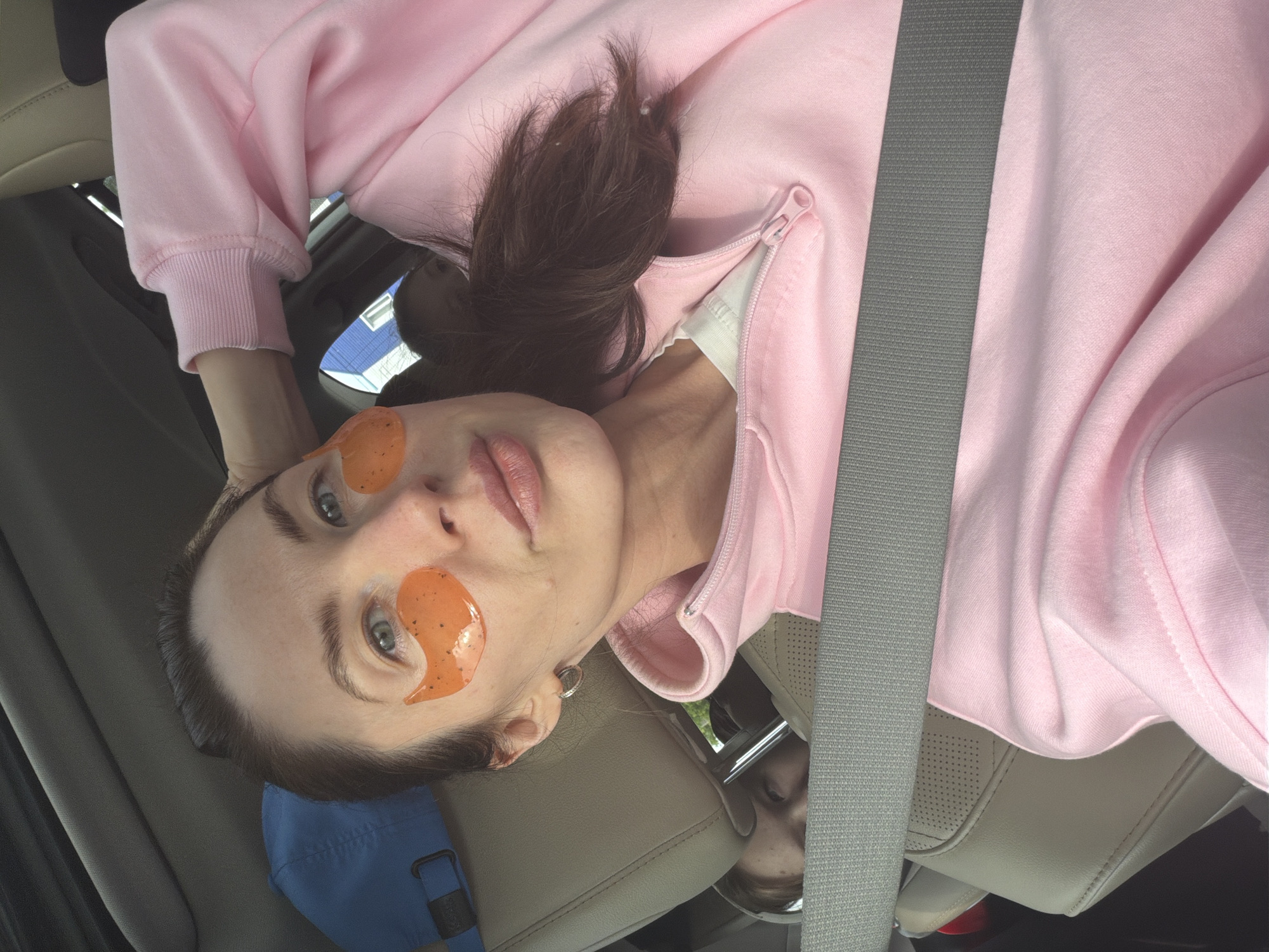 A woman is captured in a close-up, selfie-style photograph, positioned inside a car in Chaikovsky, Russia. She is wearing a light pink hooded sweatshirt with a visible white undershirt beneath the zippered neck. Her dark brown hair is loosely tied back, with some strands visible around her head. Orange, gel-like under-eye patches with small black specks are applied beneath her light blue-grey eyes, indicating a self-care activity. Her lips are slightly parted and appear natural or lightly colored. A small silver hoop earring is visible in her right ear.
She is secured by a grey car seatbelt that diagonally crosses her chest. The car interior features light tan or beige leather seats, some with perforated detailing. A blue fabric item, possibly a cap or bag, is visible near her head. Through the car window, a glimpse of a blue building with white window frames is visible in the background. Below her, partially reflected in a surface, a child's face can be seen looking upwards. The natural light indicates it is daytime. The woman's expression is calm and relaxed, conveying a sense of ease while engaged in her routine inside the vehicle.
