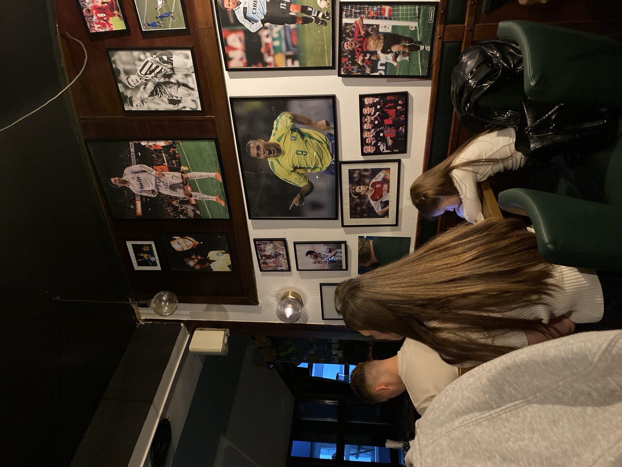 Inside a dimly lit space, likely a pub or restaurant, in Novi Sad, Serbia, two women are seated. One woman, in a white long-sleeved shirt, is using her phone, while the other, with long brown hair, is looking down. A man with short, cropped hair is also present. The walls are decorated with framed photographs of soccer players. Dark green seating is visible, along with a black leather jacket draped over a chair. A lightbulb fixture and a sconce adorn the wall. A window is seen in the background.