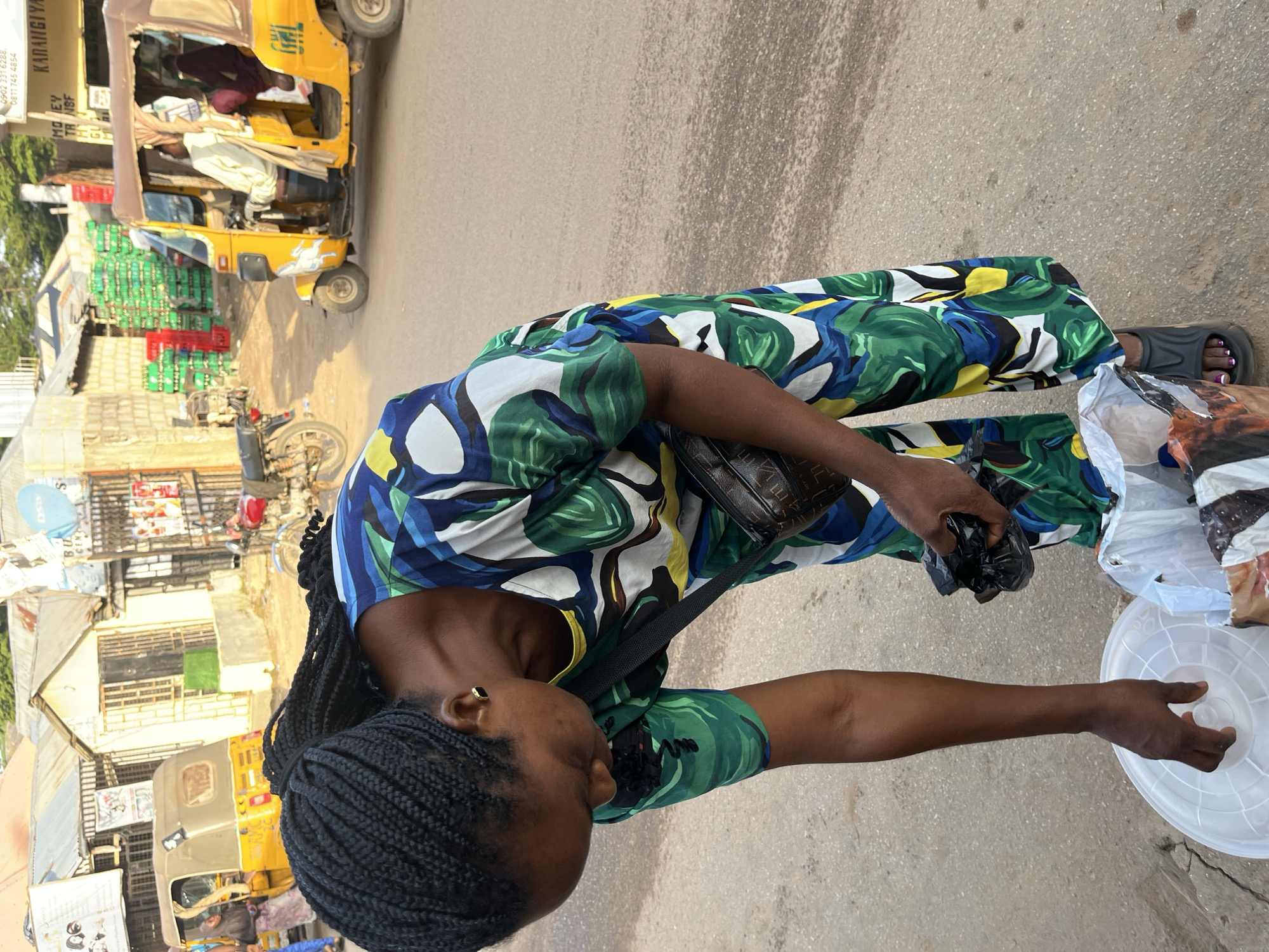 In a street scene in Abuja, Nigeria, a Black woman wearing a vibrant green, blue, and yellow patterned dress and dark braided hair is crouched down. She is holding black plastic bags in her right hand and reaching towards a white circular object with her left hand. A dark brown bag with a monogram pattern is slung across her body. The background reveals a busy street with a yellow auto-rickshaw vehicle containing passengers, a motorcycle, and buildings that appear to be small shops or stalls. Stacks of green crates, possibly containing beverages, are visible in front of one of the buildings. The ground is paved with asphalt and dirt. The lighting suggests it is daytime.