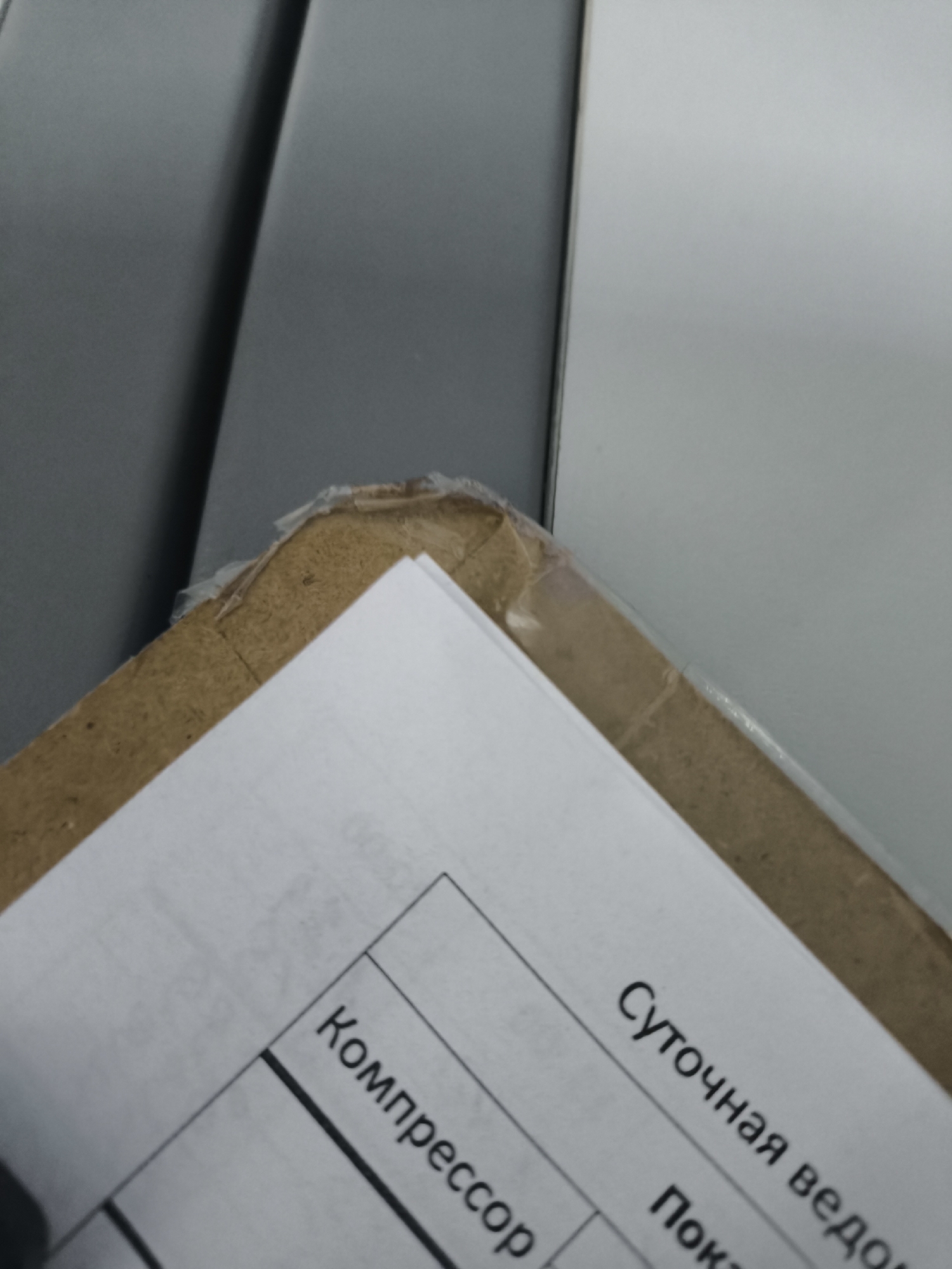 The image shows a close-up view of a document with Russian text on it, partially obscured by a cardboard edge. The document appears to be a form or report, with handwritten or printed text. The text visible includes "Компрессор" (Compressor) and "Суточная ведомость" (Daily statement). The cardboard is torn and appears to be part of a package. The background consists of a gray surface and a white surface, suggesting an indoor environment. There are no people or specific locations identifiable in the image.