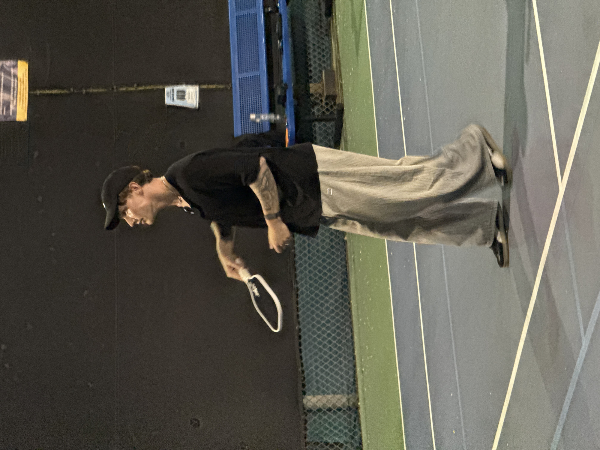 The image depicts a person standing on a sports court, likely for tennis or pickleball, at night. The subject is an adult male, captured in a full-length shot. He wears a black baseball cap, a black short-sleeved t-shirt, and wide-leg light grey pants with vertical ribbing or stripes. Tattoos are visible on his left arm. He holds a white-framed paddle or short-handled racket in his right hand. His head is turned to the left, suggesting he is looking forward or observing something off-frame, with a focused or neutral expression.
The court features distinct sections of blue and green with white boundary lines. In the background, a dark wall on the left side of the frame displays two pieces of paper or signs, though their text is illegible. To the right of the person, a chain-link fence separates the playing area from a blue metal structure, which could be part of bleachers or a facility support. A water bottle is visible resting on this blue structure.
The scene is illuminated by artificial light, indicating nighttime. The court surface and the fence show some reflective spots, possibly due to dampness or specific lighting conditions. The person's attire and the presence of the paddle suggest an activity related to racket sports. The overall setting is consistent with a recreational sports facility, located in West Hollywood, United States of America.