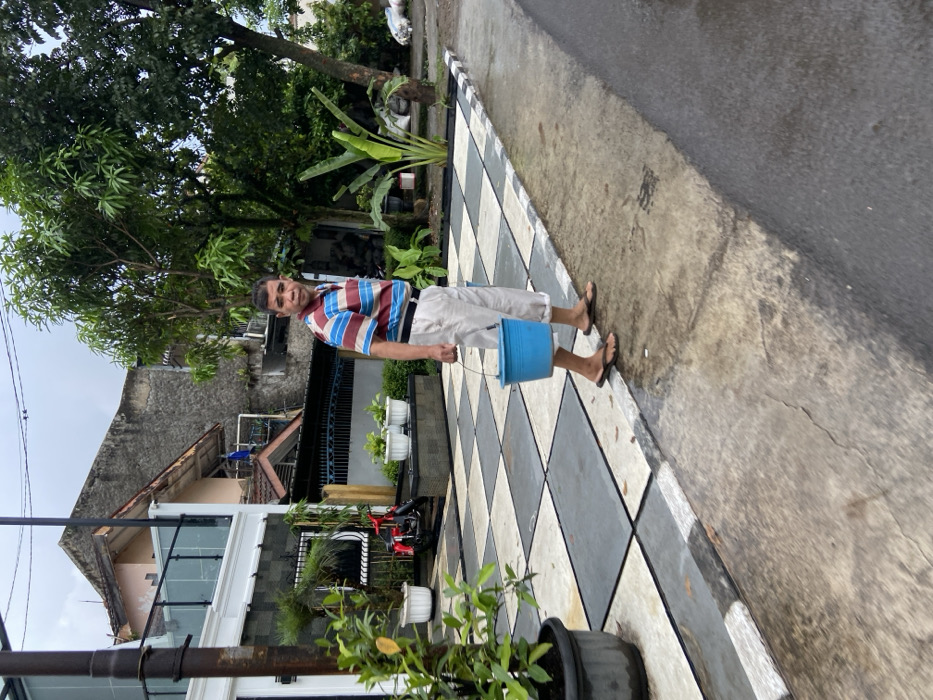 A man in a striped shirt and khaki pants is walking on a checkered concrete path towards the camera. He carries a blue bucket in his right hand. There is a motorcycle parked in the background. The path leads to a street with a black asphalt surface, the street appears to be wet. The surrounding environment is a residential area in Margaasih, Indonesia, with houses, trees, and plants. The latitude and longitude of the scene are -6.944 and 107.555, respectively.