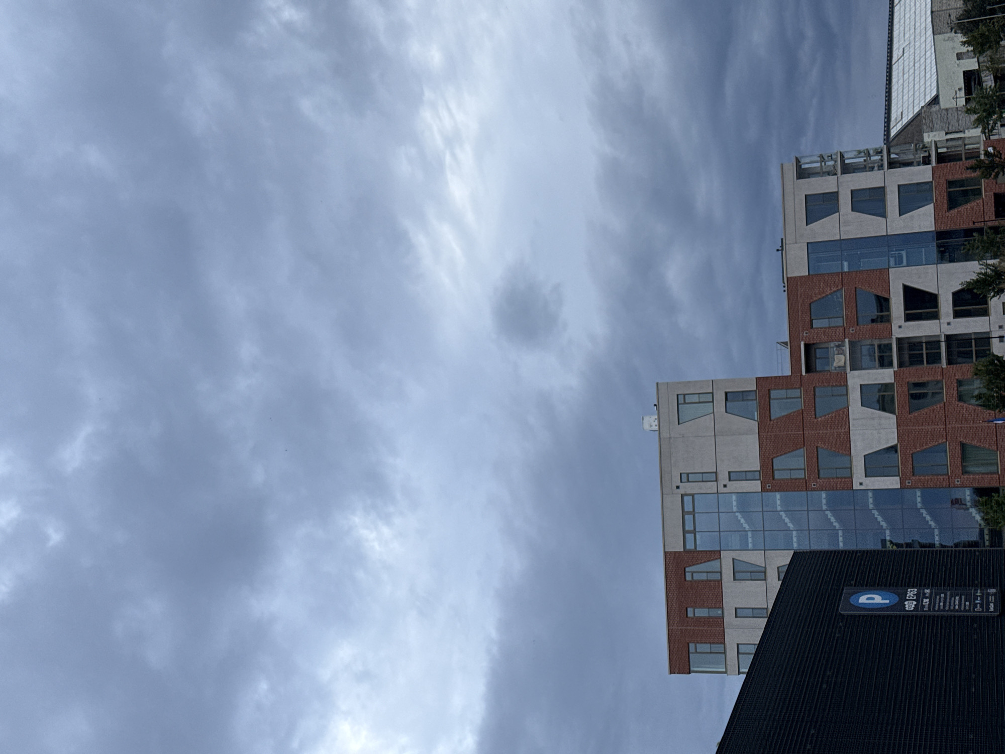The image depicts a cloudy sky over a modern building in Tallinn, Estonia. The building features a multi-colored facade with sections of red brick and white concrete, punctuated by windows of various shapes and sizes. A glass-enclosed staircase is visible in the center of the building. To the left of the main building, a dark-colored structure with a textured surface is visible, displaying a blue parking sign with a white "P" symbol and text in an unknown language. In the background, other structures and trees can be seen, along with a white roof of another building. The overall scene suggests an urban environment under overcast conditions.