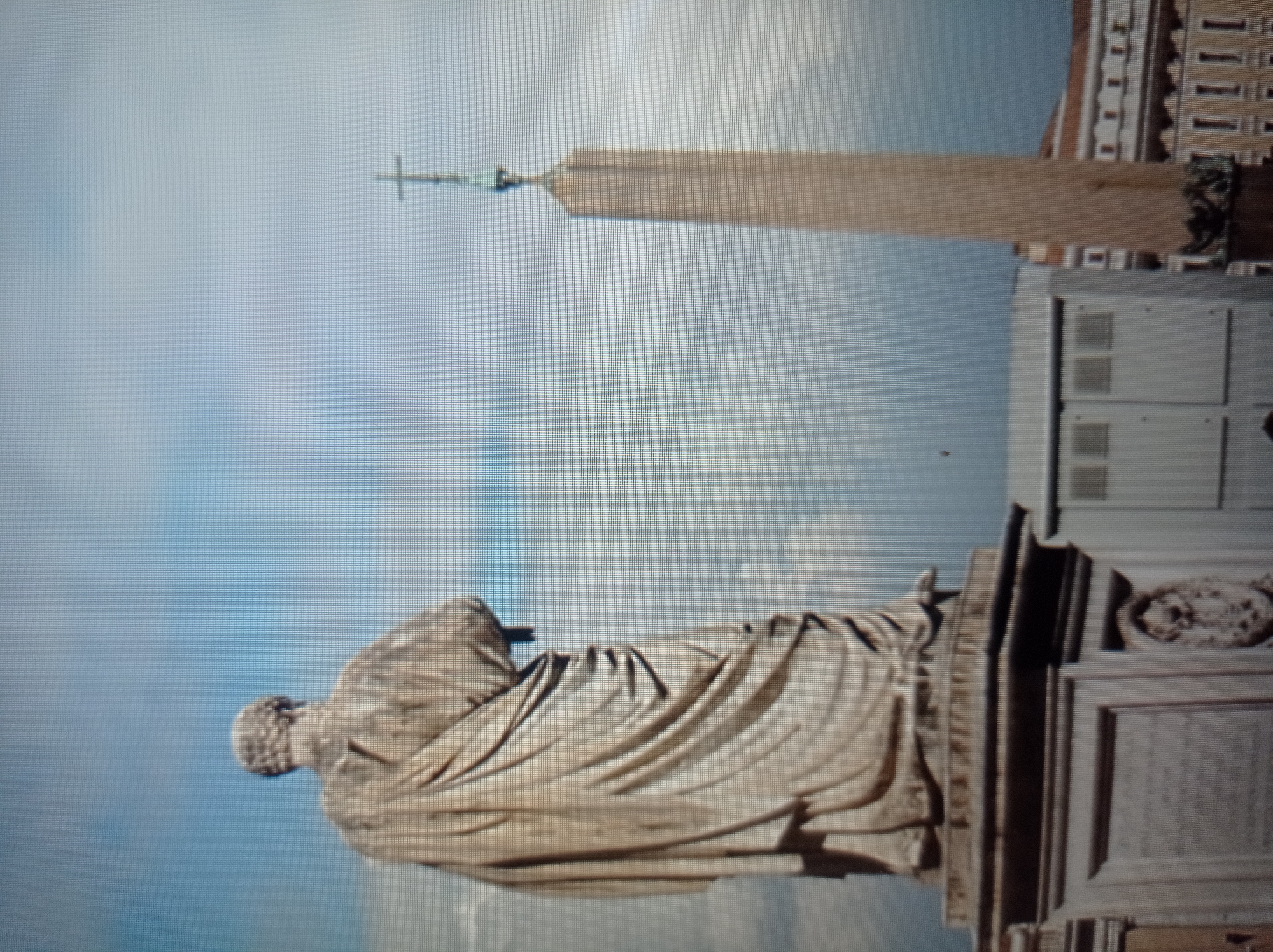 The image shows a statue of a robed figure viewed from behind, with an obelisk topped by a cross to the right. The sky is cloudy and blue. Buildings are visible in the background. The scene is likely outdoors in an urban environment, given the presence of a statue and obelisk, which are common public monuments. However, the request to include "the location in the city of Mukachevo, Ukraine" cannot be fulfilled as the provided image does not contain any visual information to confirm this specific location.
