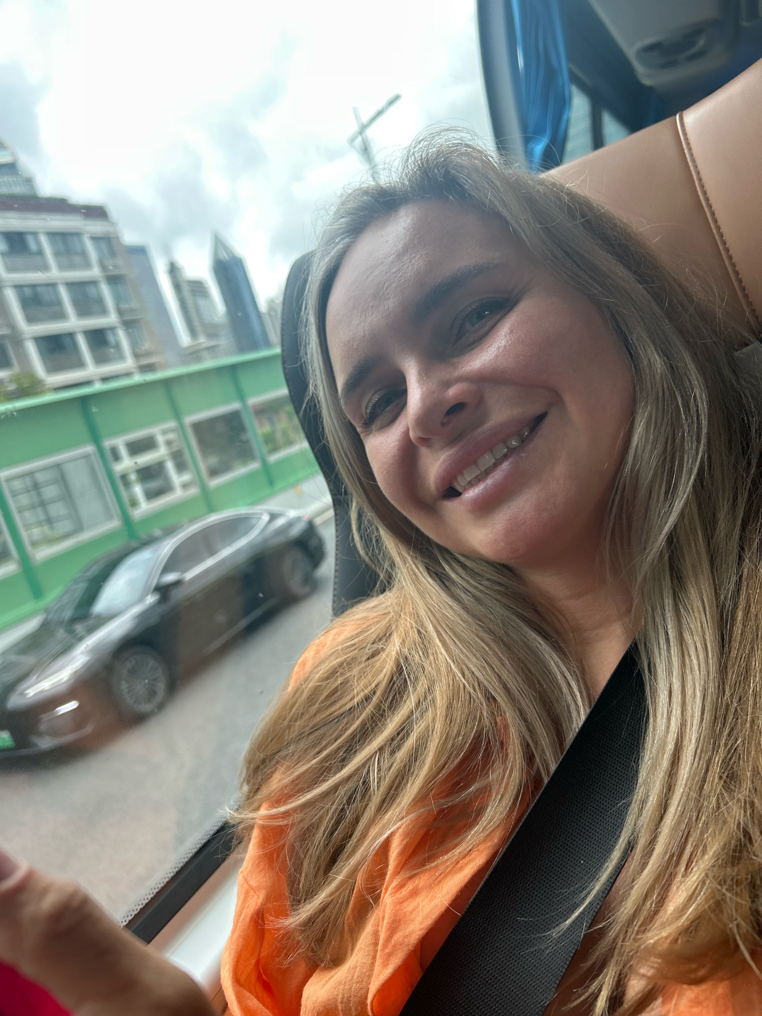 A woman with blonde hair is looking at the camera and smiling, with her mouth open. She is wearing an orange top and a black seatbelt. In the background, a black car is driving past a building with green panels. The scene appears to be from a moving vehicle, possibly a bus, with a window showing the city landscape of Jing'an District, China. The sky is overcast.