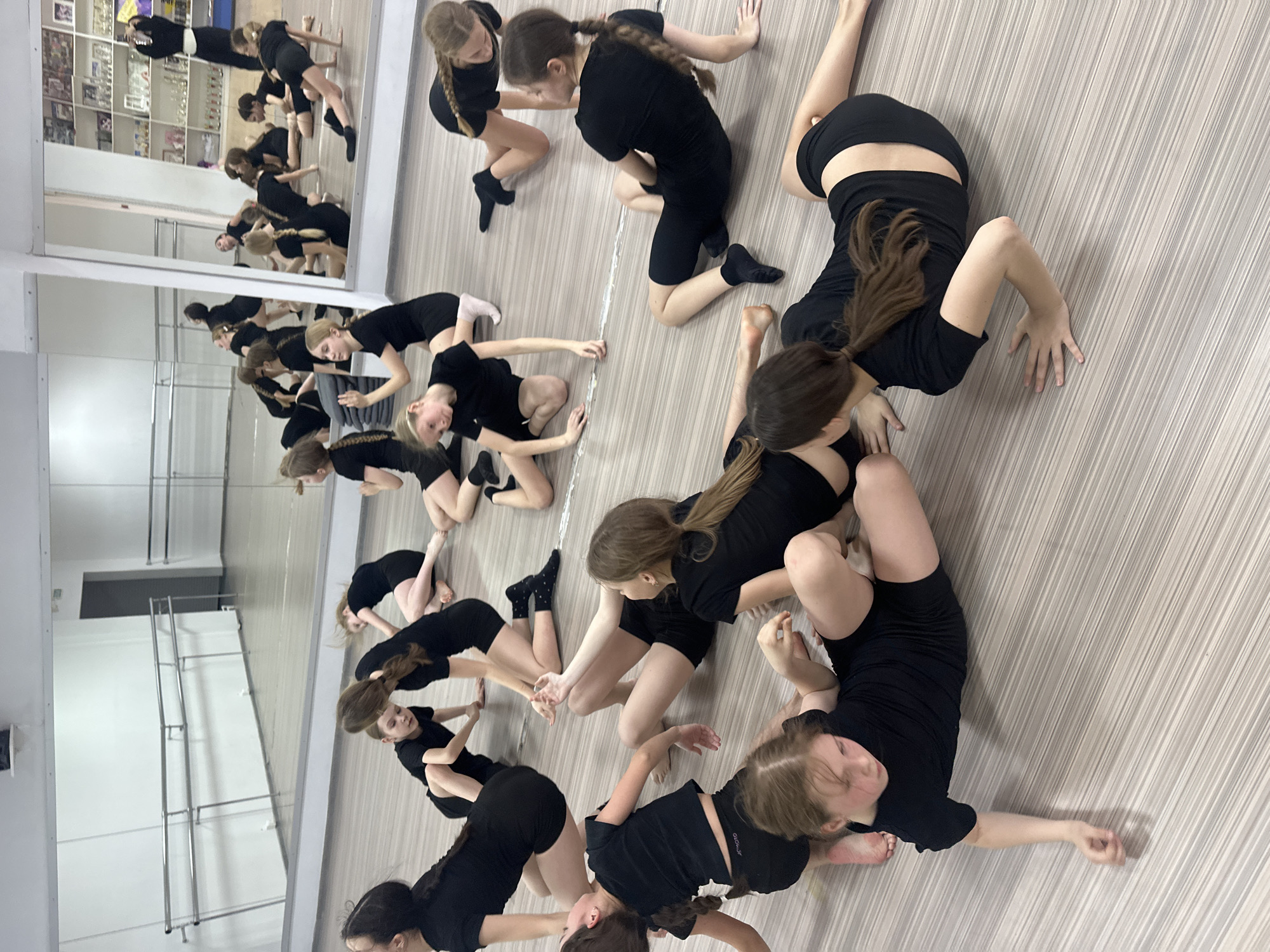 This image depicts a dynamic scene inside a dance studio in Votkinsk, Russia, showcasing a group of young females engaged in a contemporary dance or movement class. The studio features a light-colored, striped floor and a prominent mirrored wall on the left, which reflects the participants and the room's interior.
Approximately a dozen individuals, primarily children or teenagers, are visible. They are uniformly dressed in black t-shirts and black shorts or leggings, with their hair often styled in braids or ponytails. The subjects are scattered across the floor in various poses, indicative of fluid, floor-work based movements common in modern dance. Some are kneeling, others are bent forward, sitting with legs bent, or reaching out with their arms, all demonstrating concentration and controlled posture.
The mirrored wall reflects additional participants, along with ballet barres and a wall lined with shelves holding numerous trophies and awards, suggesting a long history of achievements within the studio. The perspective is slightly elevated, looking down on the dancers, emphasizing their collective engagement in the activity. The atmosphere appears focused and dedicated to practice. While small logos are visible on some of the participants' clothing, no text is clearly readable. The time of day is not explicitly indicated.