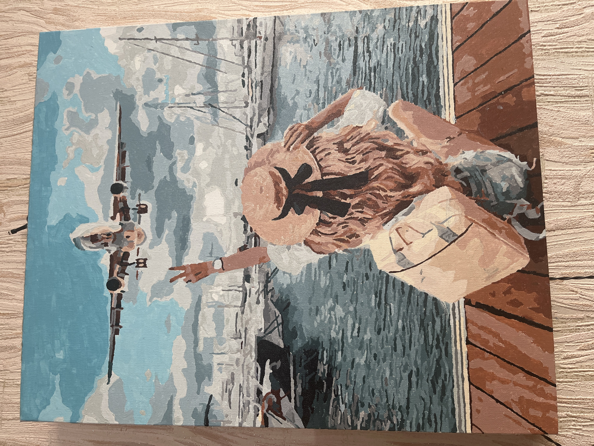 The painting depicts a young woman from behind, sitting on a wooden pier and looking out at a harbor filled with sailboats. An airplane is visible in the sky, descending towards the viewer. The woman is wearing a straw hat with a black ribbon, a white top, and denim shorts. She is holding a light-colored bag and has one hand on her hat while the other is raised, forming a peace sign. The water is a deep blue-green, and the sky is a mix of blue and white clouds. The wooden pier has a reddish-brown color with dark lines representing the gaps between the planks. The sailboats are depicted in various shades of white and gray, with tall masts. The overall style of the painting is reminiscent of a paint-by-numbers kit, with distinct areas of color and visible brush strokes. The scene evokes a sense of travel and anticipation, with the airplane suggesting arrival or departure. The specific location of Taganrog City District, Russia, is not visually discernible from the artwork itself, as the painting is a stylized representation and does not contain any identifiable landmarks or contextual clues that would place it in that specific geographical area.