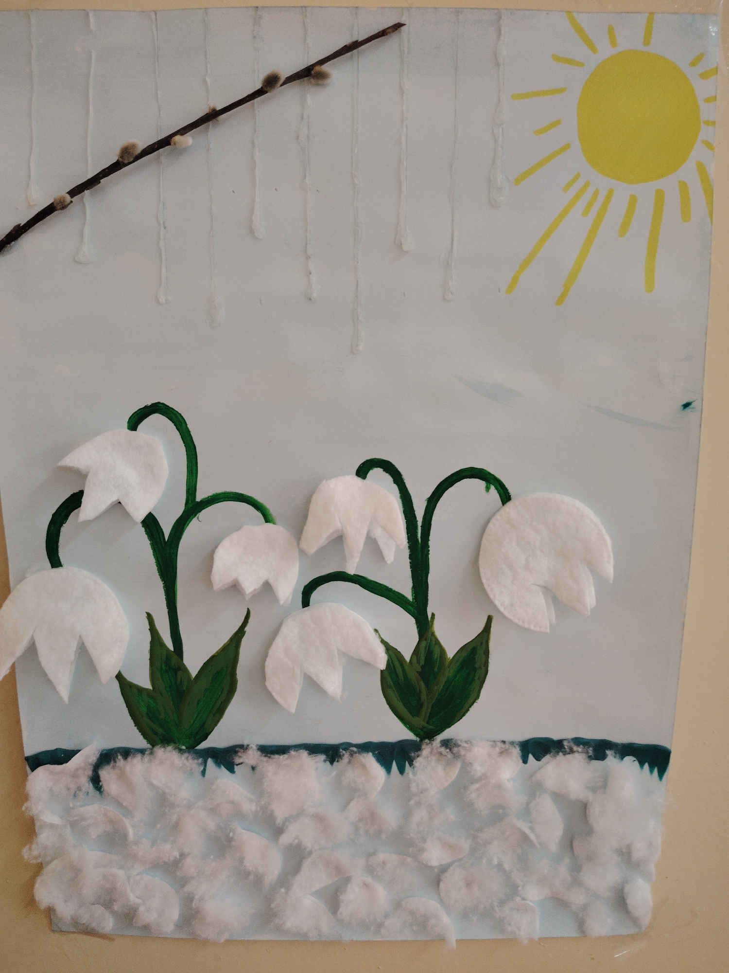 The image is a child's artwork depicting a spring scene. It features white, felt-like flowers resembling lilies of the valley with painted green stems and leaves. The flowers are set against a light blue background with a twig of pussy willow in the upper left and a yellow sun with rays in the upper right. The bottom of the artwork is covered with cotton balls, representing snow. White glue is visible in vertical drips above the flowers. There is no indication that the image is located in Sarapul, Russia.