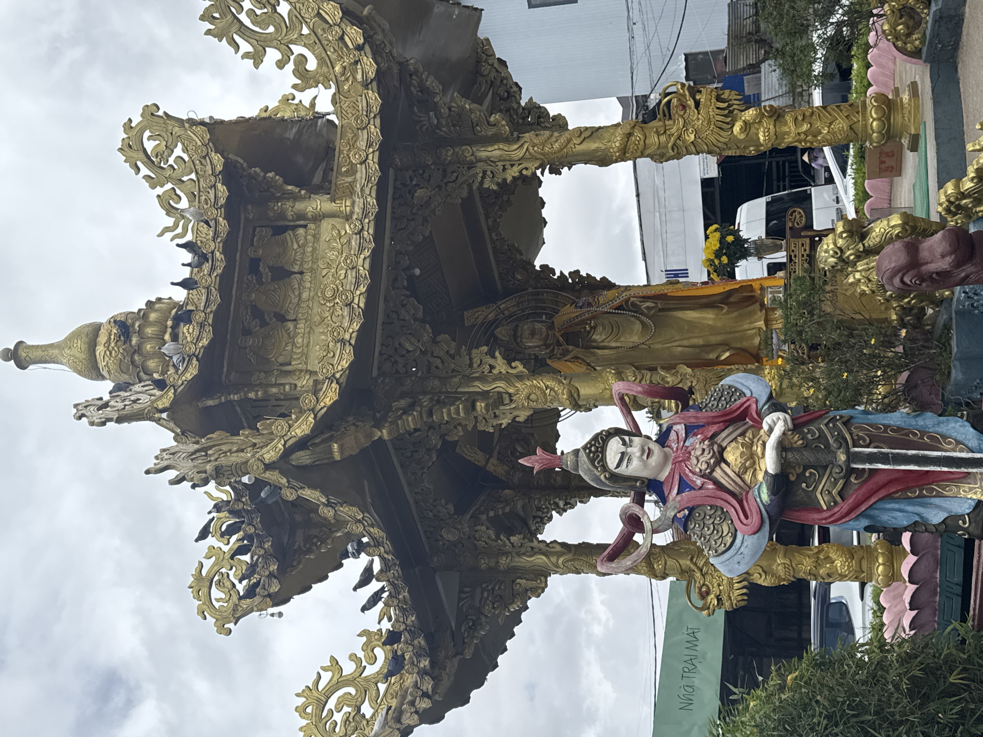 The image depicts a golden, ornate Buddhist shrine with intricate carvings, possibly in Xuân Thành, Vietnam. Pigeons are perched on the roof of the shrine. In front of the shrine stands a colorful statue of a warrior in armor, holding a sword, to the left of a golden statue of a Buddha. The surrounding environment includes lush green foliage, a lotus-shaped base, and other decorative golden dragon sculptures. In the background, buildings and vehicles are visible, suggesting an urban or semi-urban setting. A green sign with Vietnamese text is partially visible on the left. The sky is overcast.