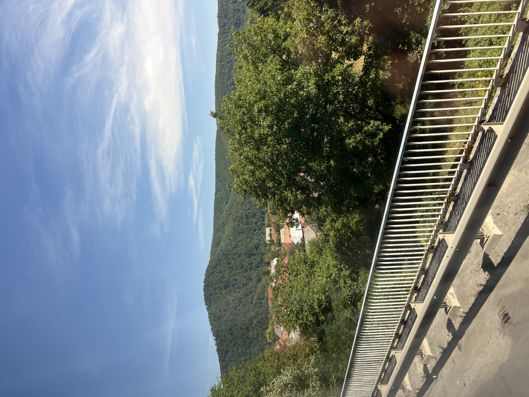 The image depicts a scenic view from a bridge overlooking the village of Chynadiiovo, Ukraine. The foreground features a metal guardrail and a concrete surface, likely part of the bridge. Beyond the bridge, a cluster of houses with red-tiled roofs is visible, nestled amongst lush green trees. In the background, a heavily forested hill rises, dominating the landscape. The sky is a clear blue with wispy white clouds, suggesting fair weather. The overall impression is one of a peaceful, rural setting.
