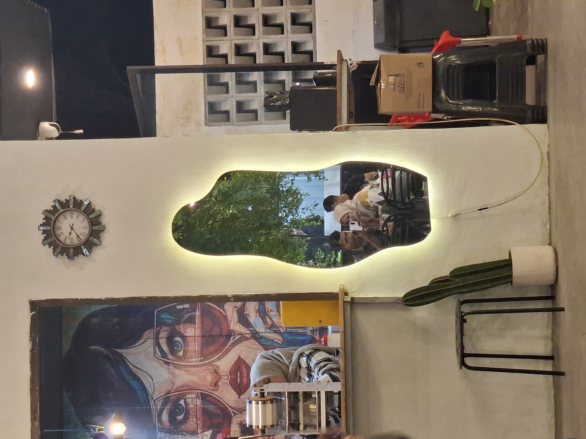 The image captures an interior scene, likely in Lhokseumawe, Indonesia. A large, irregular-shaped mirror with a glowing edge reflects a scene with people sitting and possibly interacting at a table, surrounded by greenery. Below the mirror, a framed artwork depicting a woman with large glasses dominates the wall space.
To the left of the mirror is a decorative clock. To the right is a cactus in a white pot placed on a black metal stand. In the background, a wall with a textured geometric pattern is visible. Various items, including boxes and what appears to be cleaning equipment, are stacked behind the mirror. The overall lighting suggests an evening setting.