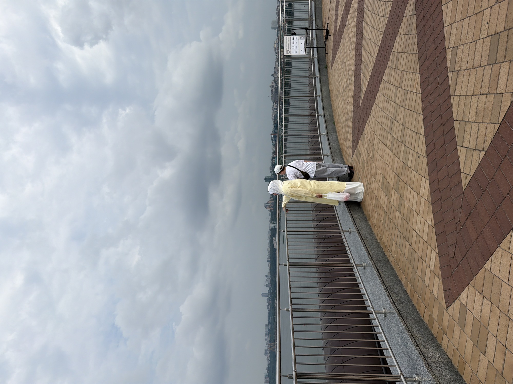 Two individuals stand near a metal railing, overlooking a wide body of water with a cityscape on the opposite shore. The person on the left wears a yellow abaya and a white hijab, carrying a white plastic bag. To their right, another person is dressed in a white t-shirt and grey pants, wearing a white cap. The foreground features a paved walkway with a pattern of beige and brown rectangular tiles. The sky is overcast with heavy, grey clouds. The location is identified as a viewing area in the city of Sapogoli, Russia, suggested by a sign in Russian visible on the right side of the image.
