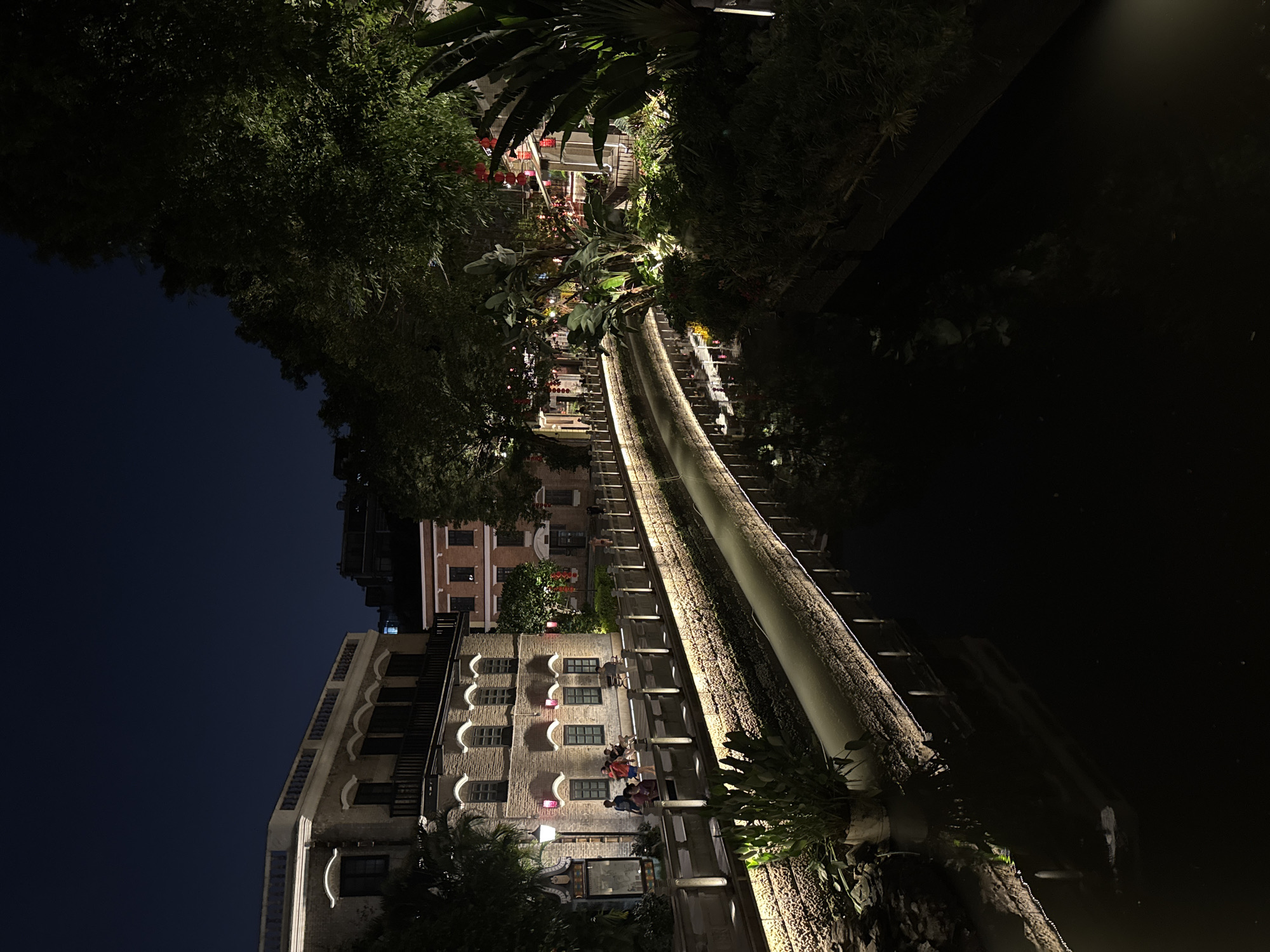 The image shows a nighttime view of a waterway lined with buildings in the city of Liwan District, China. The buildings are illuminated with various lights and feature ornate architectural details, including arched windows and balconies. People can be seen on some of the balconies. The waterway reflects the lights of the buildings, creating a mirrored effect. Lush trees and foliage surround the waterway, adding a natural element to the scene. The sky above is dark.