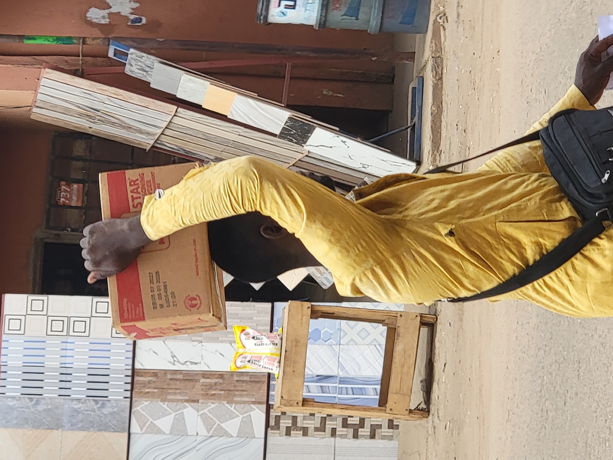 A man wearing a mustard-yellow shirt carries a cardboard box on his head. He is also wearing a black bag slung across his body. The man is walking on a street or sidewalk next to a building that sells tiles. Various tiles are displayed for sale, including designs with squares, geometric patterns, and wood-like textures. A stack of wooden tiles leans against the building. The setting is in a city, likely Maiburiji, Nigeria.