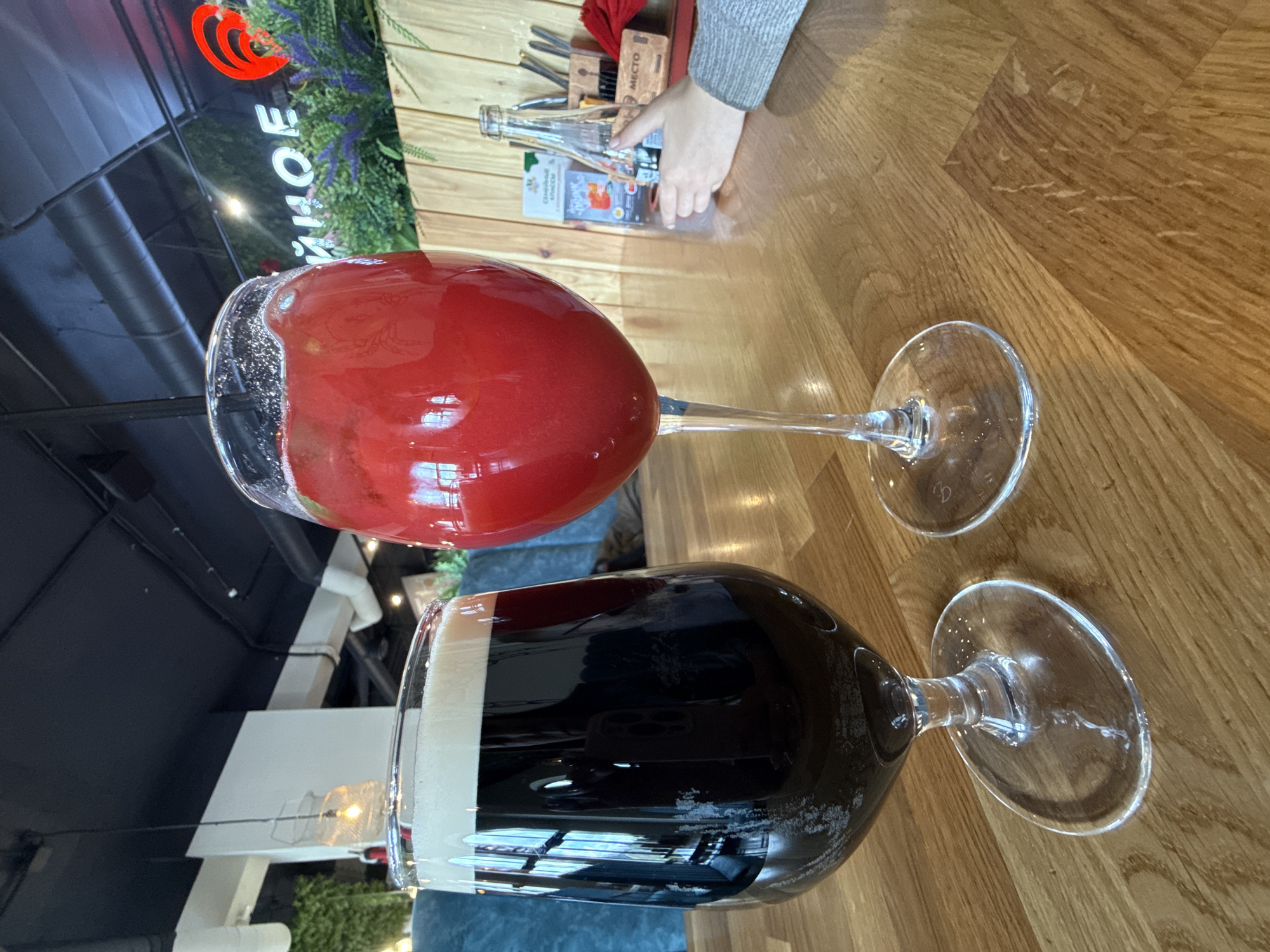 Two filled wine glasses are resting on a wooden table. One glass is filled with a red liquid, possibly wine or a cocktail, while the other contains a dark liquid, likely beer or stout, with a foamy head. In the background, a hand is visible near cutlery and bottled drinks. A sign with "ZHOE" in Cyrillic script is seen, possibly indicating the name of the establishment. The image was taken in Dmitrov, Russia.