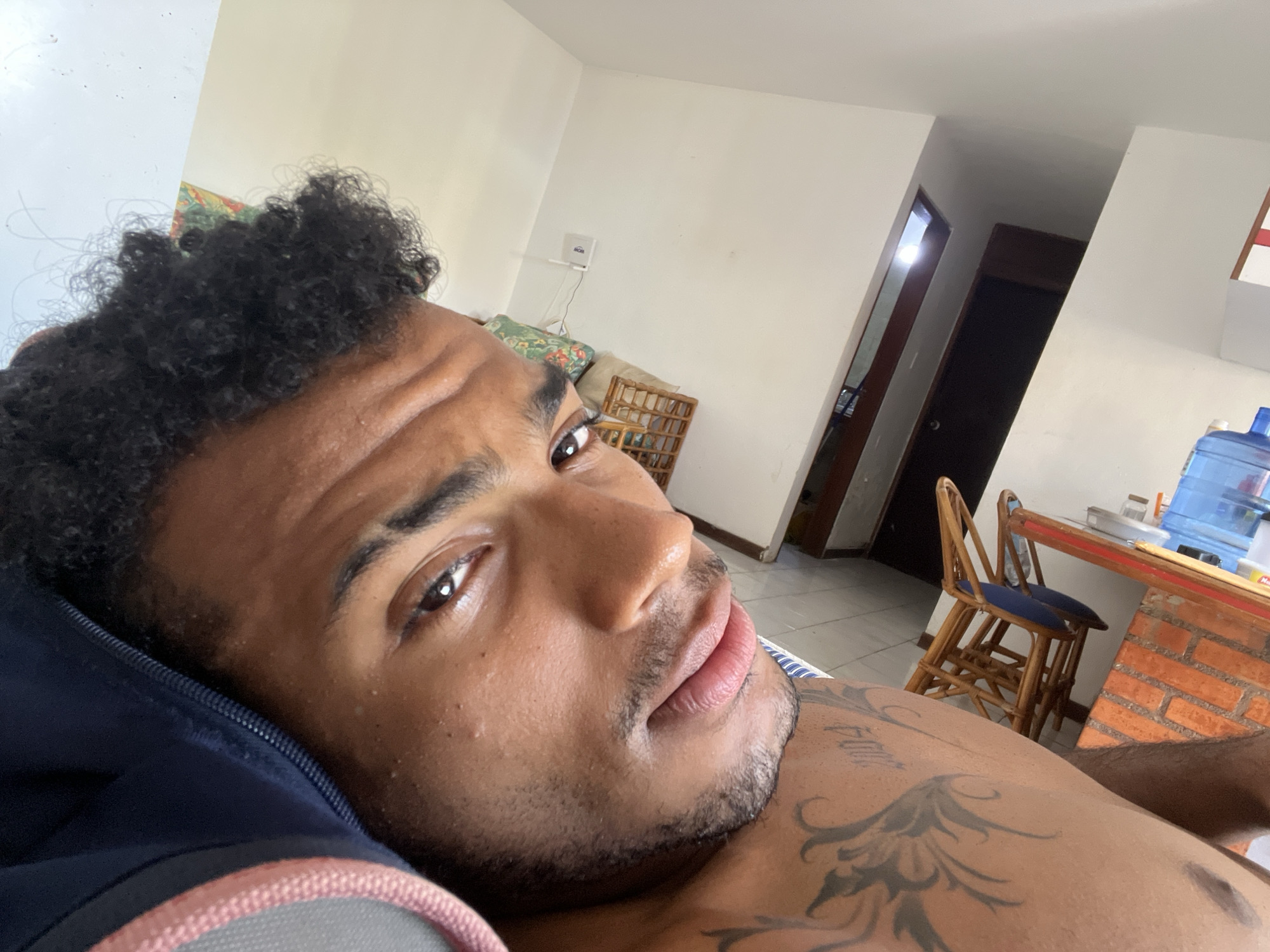 A close-up, tilted selfie of a light-skinned man with short, curly black hair. He is lying down, with part of a dark blue and grey pillow visible on his left. He has dark eyes, a trimmed beard, and a tattoo of a floral design on his chest. The background is an interior space with white walls, tiled floors, and an open doorway. Visible furniture includes a wooden chair with a blue cushion, and a red brick counter with a water cooler jug on top. A white wireless router is mounted on the wall. The location cannot be pinpointed in Porlamar, Venezuela, with the available visual data.