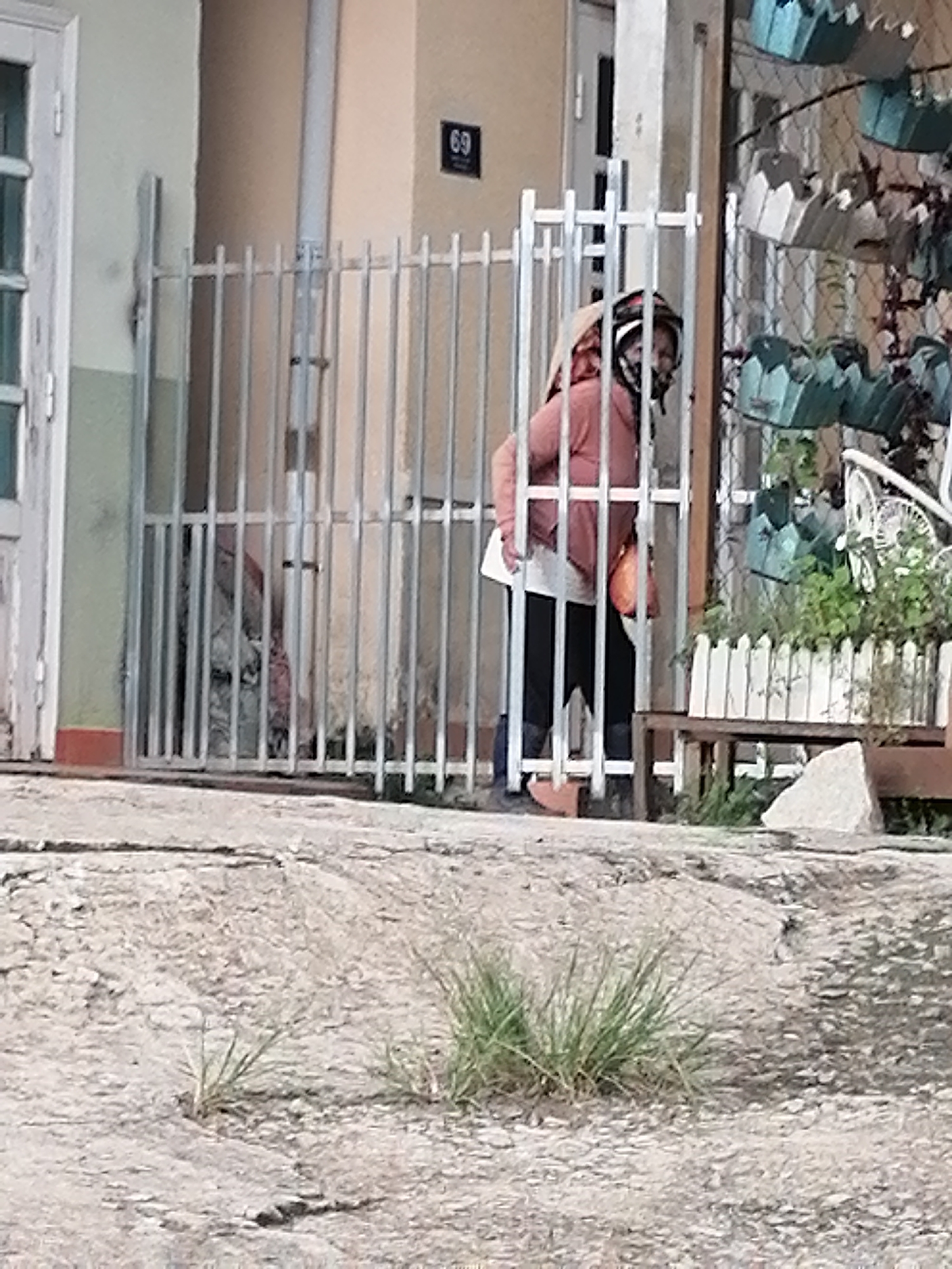 A person wearing a pink hoodie and black pants is standing behind a metal gate in Xuân Thành, Vietnam. The person is wearing a headscarf and a mask. In the background, there is a house with a number 69 on the wall and hanging planters. The ground is paved and there is some dry grass growing in cracks.
