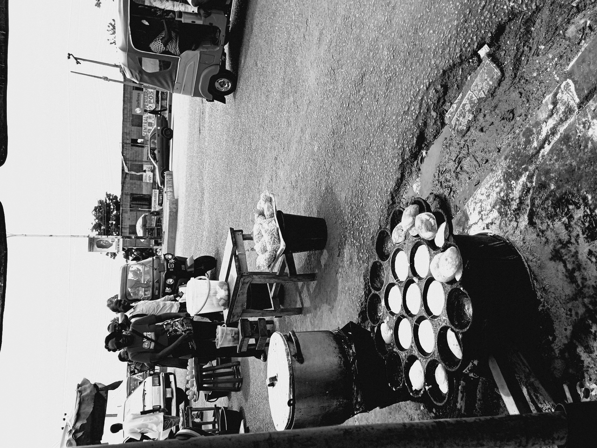 The image depicts a street scene in Bukuru, Nigeria. A vendor is preparing and selling food from a roadside setup. The food, likely "masa," is being cooked in a specialized pan with multiple circular molds. Buckets of the uncooked batter sit nearby on a chair. Customers are seen standing near the food stall, presumably waiting to purchase the food. Cars and auto rickshaws (tuk-tuks) are also visible, which indicates the stall is on a busy street in Bukuru. The surface around the cooking area is dirty and unpaved, with evidence of spilled liquids and mud. A sign for StarTimes and a supermarket are in the background.