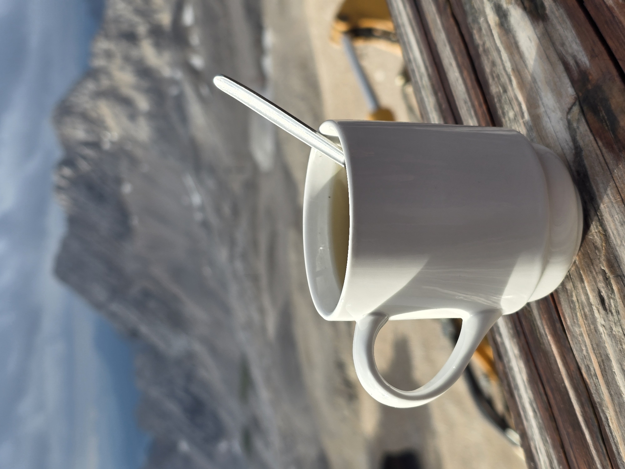 A white ceramic mug filled with a light-colored liquid, possibly tea or coffee, sits on a wooden table. A silver spoon is partially submerged in the liquid. In the background, blurred mountains and a cloudy sky are visible, suggesting an outdoor, high-altitude location. The scene appears to be from a mountaintop or a scenic viewpoint, possibly in the Bavarian Alps near Garmisch-Partenkirchen, Germany, given the mountainous landscape. There are no people or animals present in the image. The lighting suggests it is daytime, with sunlight casting shadows.