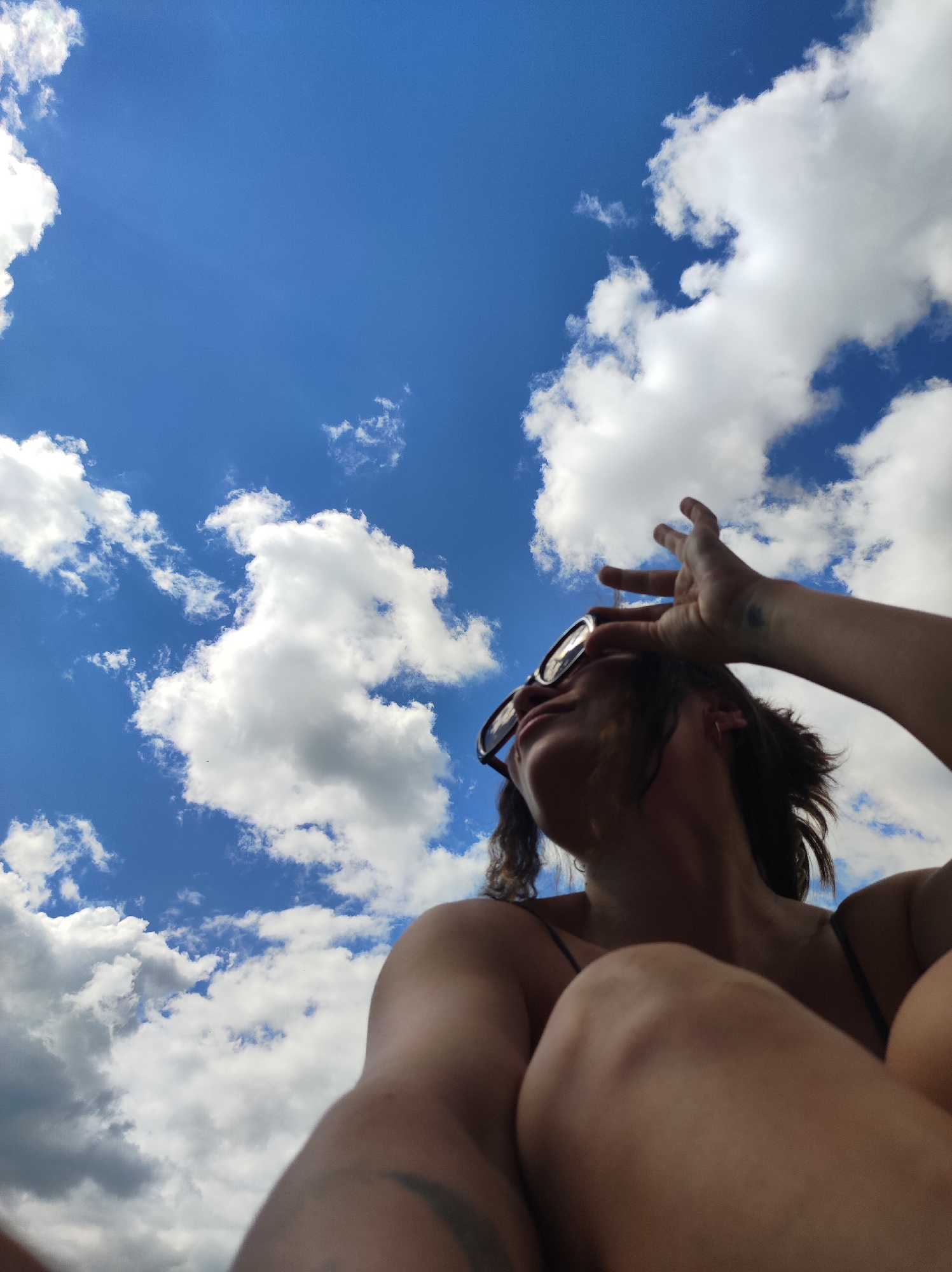 A low-angle shot captures an individual, likely female, observed from below against a bright blue sky dotted with numerous white cumulus clouds, in the city of Фоки, Russia. The person is positioned in the lower frame, looking upwards, wearing dark-framed sunglasses. Their right arm is raised, with the hand partially obscuring the upper right side of their face and temple, possibly shielding from sunlight or adjusting eyewear. A small, dark tattoo is visible on the inner wrist of the raised arm. Dark, wavy hair frames the face. The individual wears a dark-colored strap garment, possibly a tank top or swimsuit. In the lower right foreground, a bent knee and thigh are visible, indicating a seated or reclined posture. Strong sunlight illuminates the scene, creating bright highlights and some shadow on the subject.