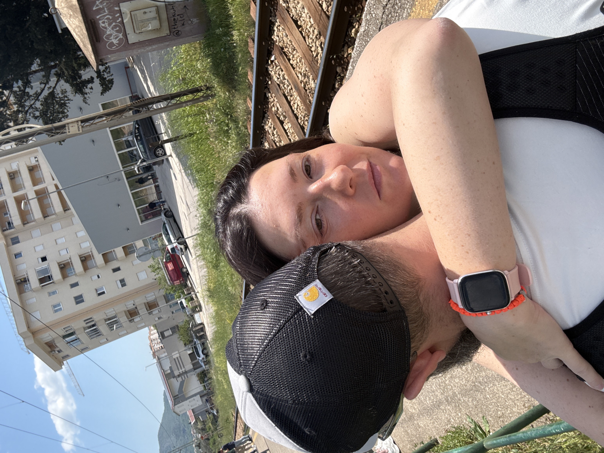 A close-up, canted-angle shot depicts a woman and a man lying together outdoors in Bar, Montenegro. The woman, with dark hair, lies facing the camera, her gaze directed at the lens. The man, wearing a black and white Carhartt hat, is partially visible behind her, his face turned away. The woman wears a smartwatch with a pink band and an orange decorative bracelet on her left wrist. They are positioned next to a set of railroad tracks. Background details include a multi-story building and parked cars on the left, with green vegetation visible along the edges of the scene. The lighting suggests a sunny day.