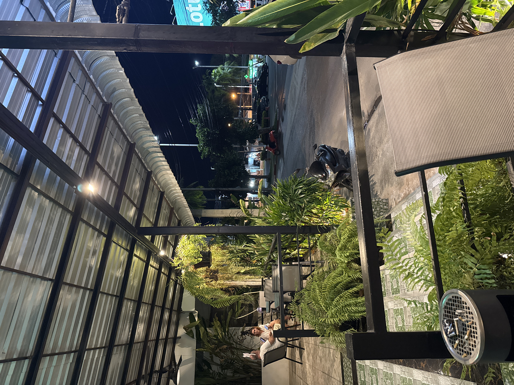 An exterior shot taken at night shows a covered patio area of a restaurant or cafe. A corrugated metal roof is supported by dark metal beams and posts. Several pendant lights hang from the ceiling, and the area is illuminated by artificial lighting. In the foreground, a gray metal chair with a striped seat is positioned in front of a black metal railing. To the left of the chair is an outdoor ashtray filled with cigarette butts. Beyond the railing, lush green ferns and tropical plants are visible. A couple is seated at a table on the patio, with the man wearing bunny ears. In the background, a street is visible with parked cars, motorbikes, and streetlights. A sign with "Lotus" in neon green is also visible.