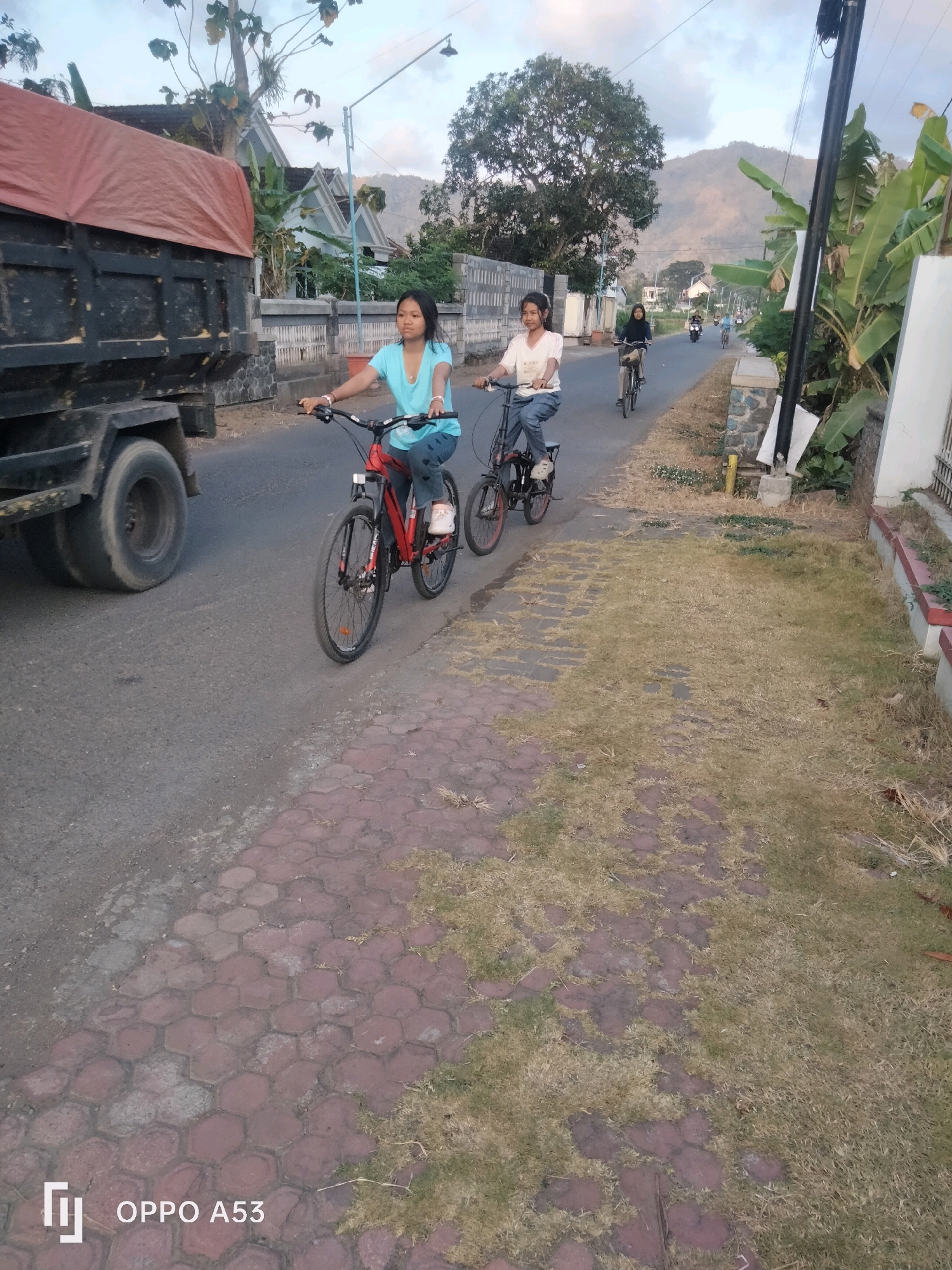 In Ngulanwetan, Indonesia, two young girls are riding bicycles on a paved road alongside a sidewalk. The girl in the foreground, wearing a blue shirt and jeans, is on a red bicycle. To her right, another girl in a white shirt and jeans rides a dark-colored bicycle. Behind them, several other individuals are also cycling, and a truck with a red tarp-covered bed is parked on the side of the road. The environment includes houses with white walls and gray roofs, a large tree, and a mountainous landscape in the background. The sky is overcast.