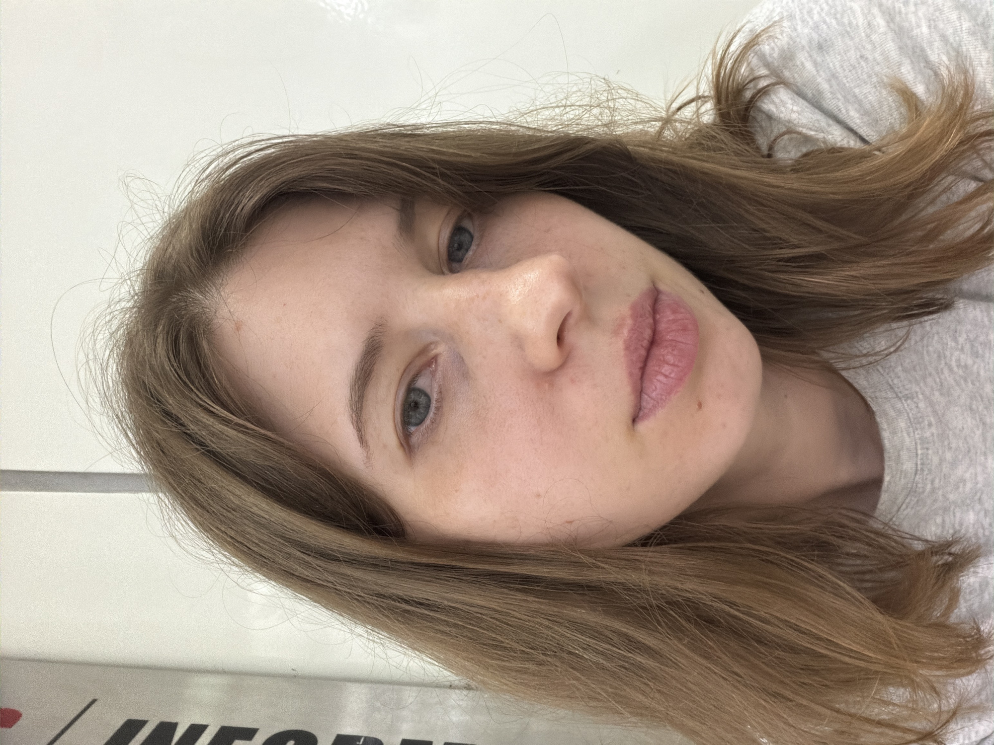 A head and shoulders, close-up, indoor portrait shows a young woman with shoulder-length brown hair and blue eyes looking forward with a neutral expression. She is wearing a grey t-shirt. The background is a plain white wall with a vertical dark stripe to the left. The image was taken on the outskirts of Seoul, South Korea.
